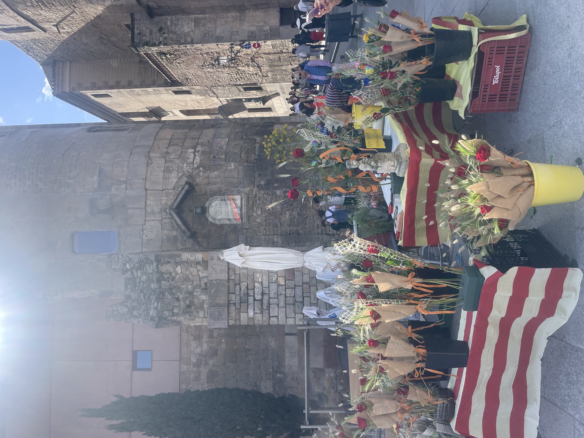 The image shows a scene in Barcelona, Spain, likely a street market celebrating Sant Jordi's Day. Several stalls display red roses wrapped in burlap and tied with ribbons, some arranged in buckets or displayed on tables draped with the Catalan flag (red and yellow stripes). A crowd of people is visible browsing the stalls, some wearing casual clothing. In the background, there's a stone building, possibly part of the Gothic Quarter, with architectural details like windows and a tower. The overall setting suggests a vibrant street market atmosphere.