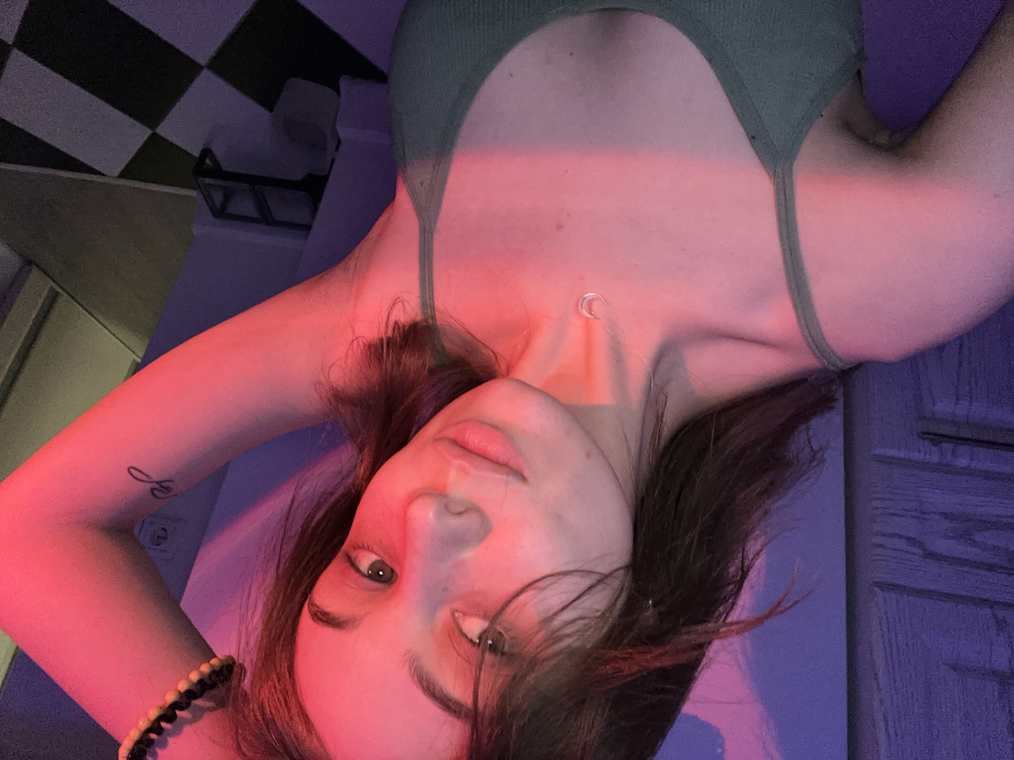 A young woman poses for a selfie. She has brown hair and wears a green tank top and a moon necklace. She is lit with pink and purple lighting, and a bracelet is visible on her arm. The background includes a checkered pattern and a door with raised panels. It is not possible to determine the location of the photo as Moscow, Russia.