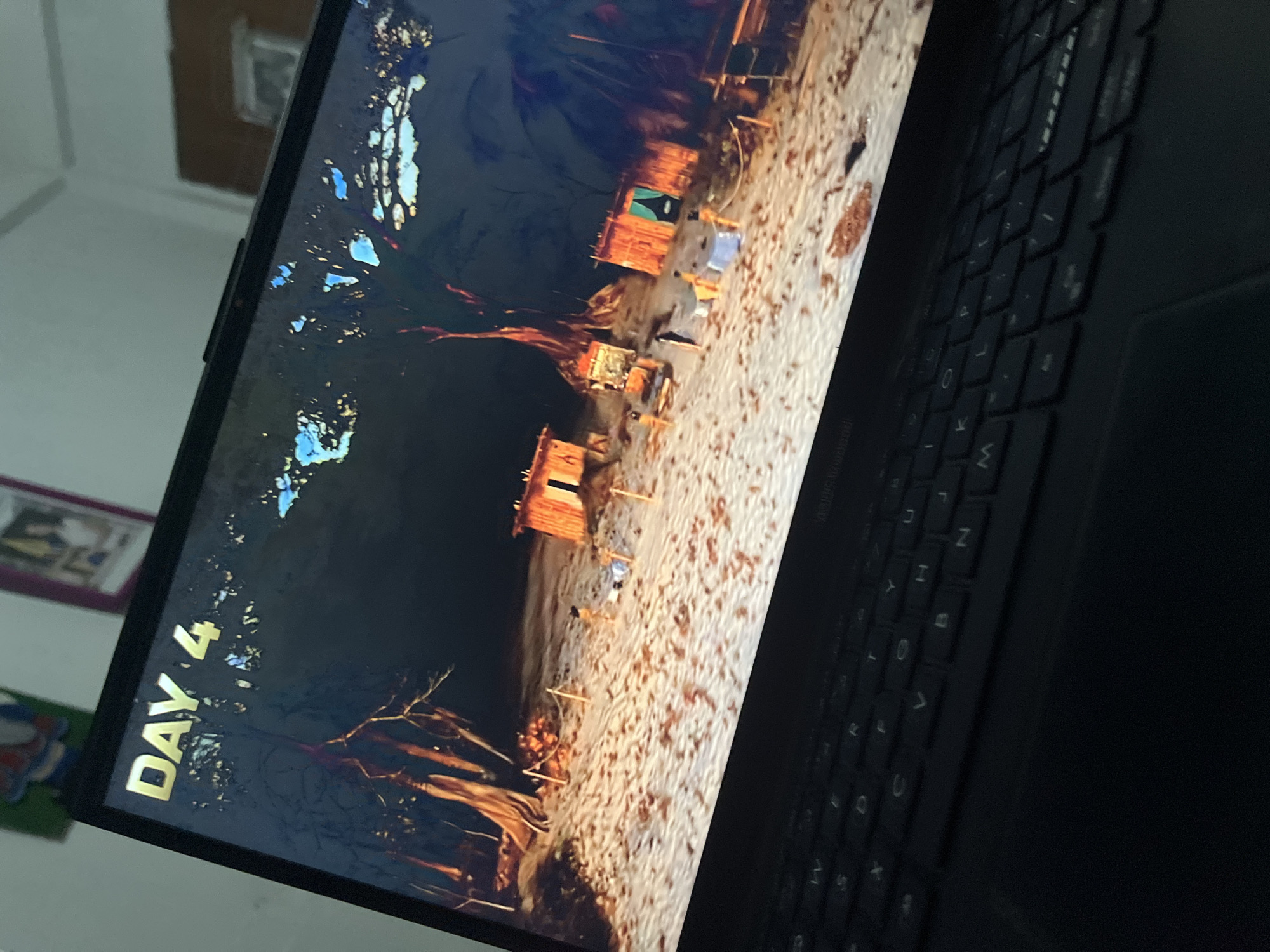 The image displays an overhead, angled view of a black laptop with its screen open. The laptop features a visible keyboard in the foreground, and the "ASUS Vivobook" branding is subtly present on the hinge area below the screen. A webcam is integrated into the top bezel of the screen.
The laptop screen displays what appears to be a digital environment from a video game or simulation. In the upper left corner, the text "DAY 4" is rendered in gold, three-dimensional letters. The scene depicts a dark environment, possibly night or dusk, with a light-colored sandy or dirt ground. Multiple tall, gnarled trees with prominent roots are visible in the background and mid-ground, suggesting a tropical or forested setting. Scattered across the sandy area are several small, rustic wooden structures resembling huts or shacks. Small, human-like figures are visible on the ground, near and between these structures, appearing to be standing or engaged in various activities. Rope fences or boundaries are staked into the ground in the foreground, and a piece of blue fabric is laid out near one of the central huts.
The area surrounding the laptop is out of focus, indicating an indoor environment. A light-colored wall is visible in the background, along with a blurred framed picture or artwork and a partial view of what may be a ceiling fan or light fixture. The image provides no identifiable geographical markers to determine a specific city, such as Santa Ana, Venezuela, or any other location.