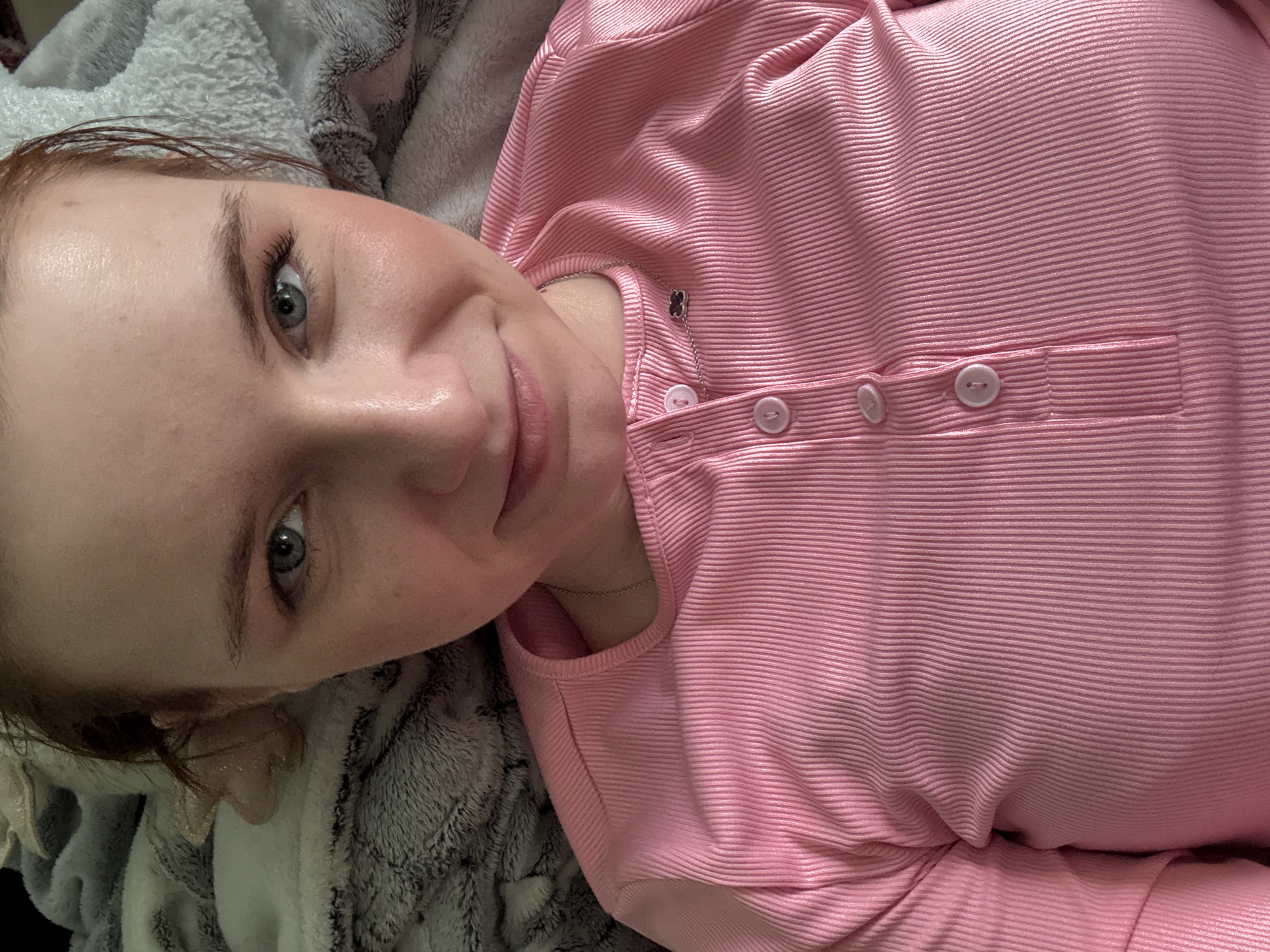 A pale-skinned woman with light blue eyes and reddish-brown hair is lying down on a gray and white patterned blanket. She is wearing a pink, ribbed long-sleeved shirt with four white buttons. A silver necklace with a black clover-shaped pendant is visible around her neck. The image is taken from an overhead perspective. No evidence suggests a location in Unknown, Russia.
