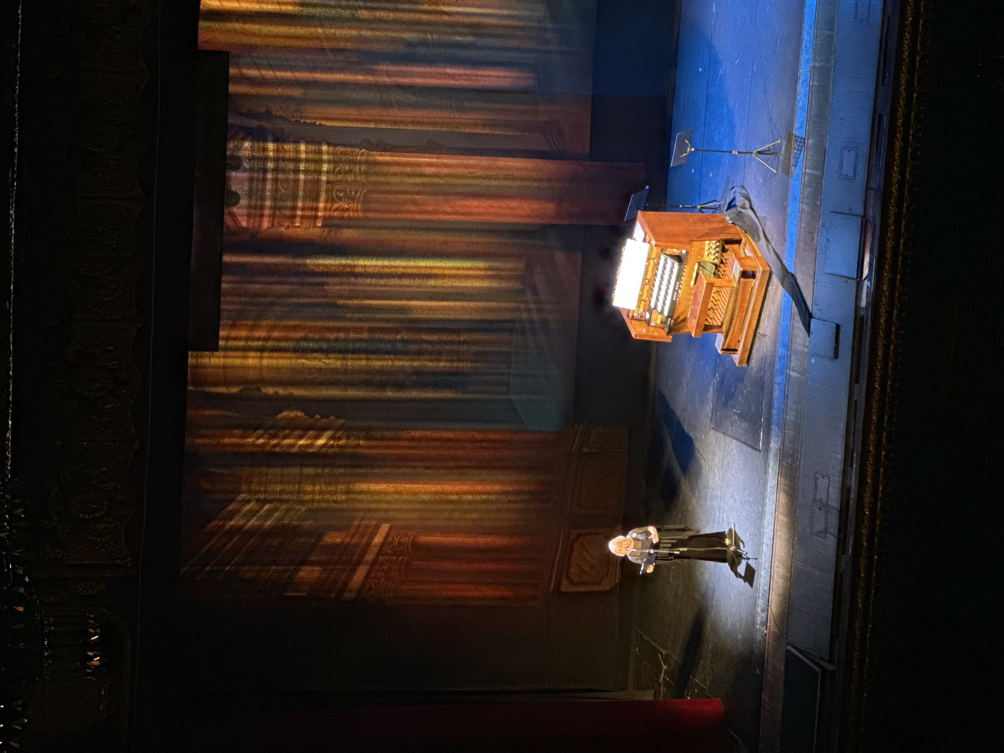 An elevated, wide shot captures a stage at the Odesa National Academic Theater of Opera and Ballet in Odesa, Ukraine. The stage is set with a backdrop resembling classical architecture, featuring tall columns illuminated by warm, vertical spotlights that create a dramatic effect. On the left side of the stage, a woman with blonde hair, wearing a black dress with fringe details, stands at a podium and speaks into a microphone. To her right, a small, wooden electronic organ is positioned on the stage. The stage floor is illuminated with blue and white lights. A red curtain is visible on the far left edge of the frame. The foreground shows the dark seating area of the theater, indicating the viewpoint is from an audience member.