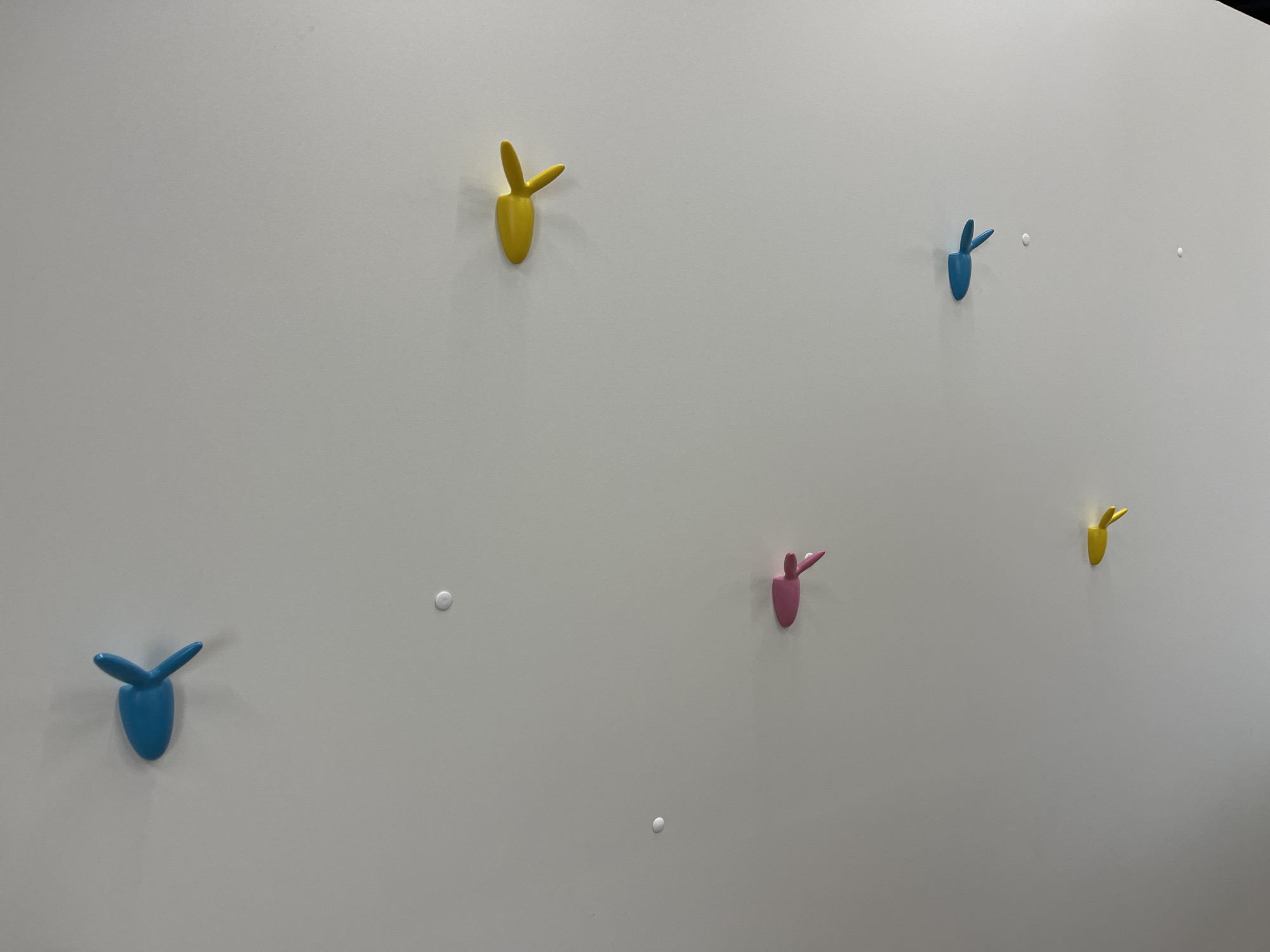 The image shows a white wall with seven colorful, stylized coat hooks attached to it. There are two yellow, two blue, and one pink hook. The hooks are spaced unevenly across the wall. Also visible on the wall are three round, white nail protectors. No people or other objects are visible in the image. There is no information given about the location.