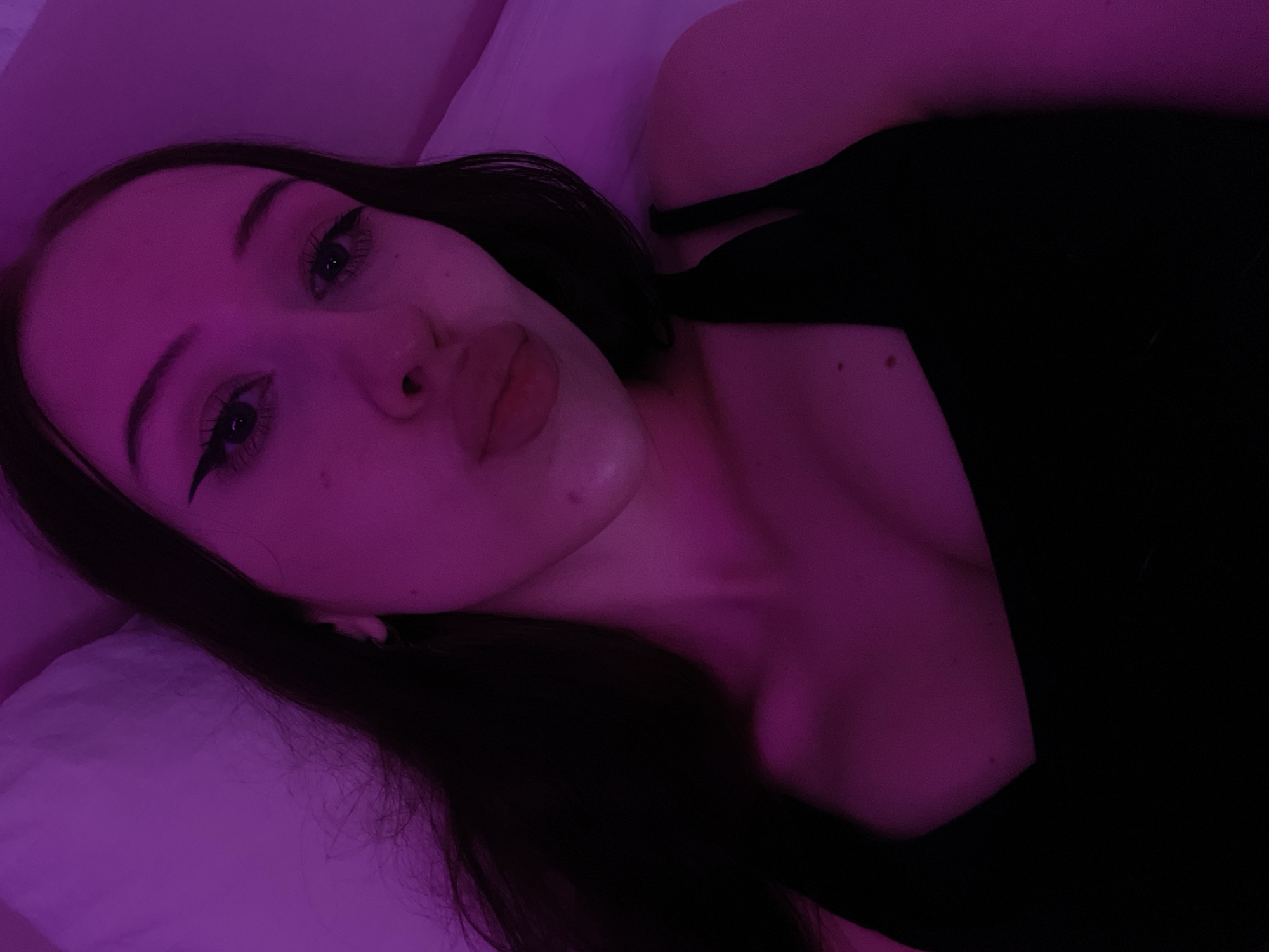 A young woman with dark hair and noticeable eyeliner is in a close-up selfie. She is wearing a black top and her lips are puckered. The scene is illuminated by purple light, and she appears to be lying on a white surface, possibly a bed. The location is Votkinsk, Russia.