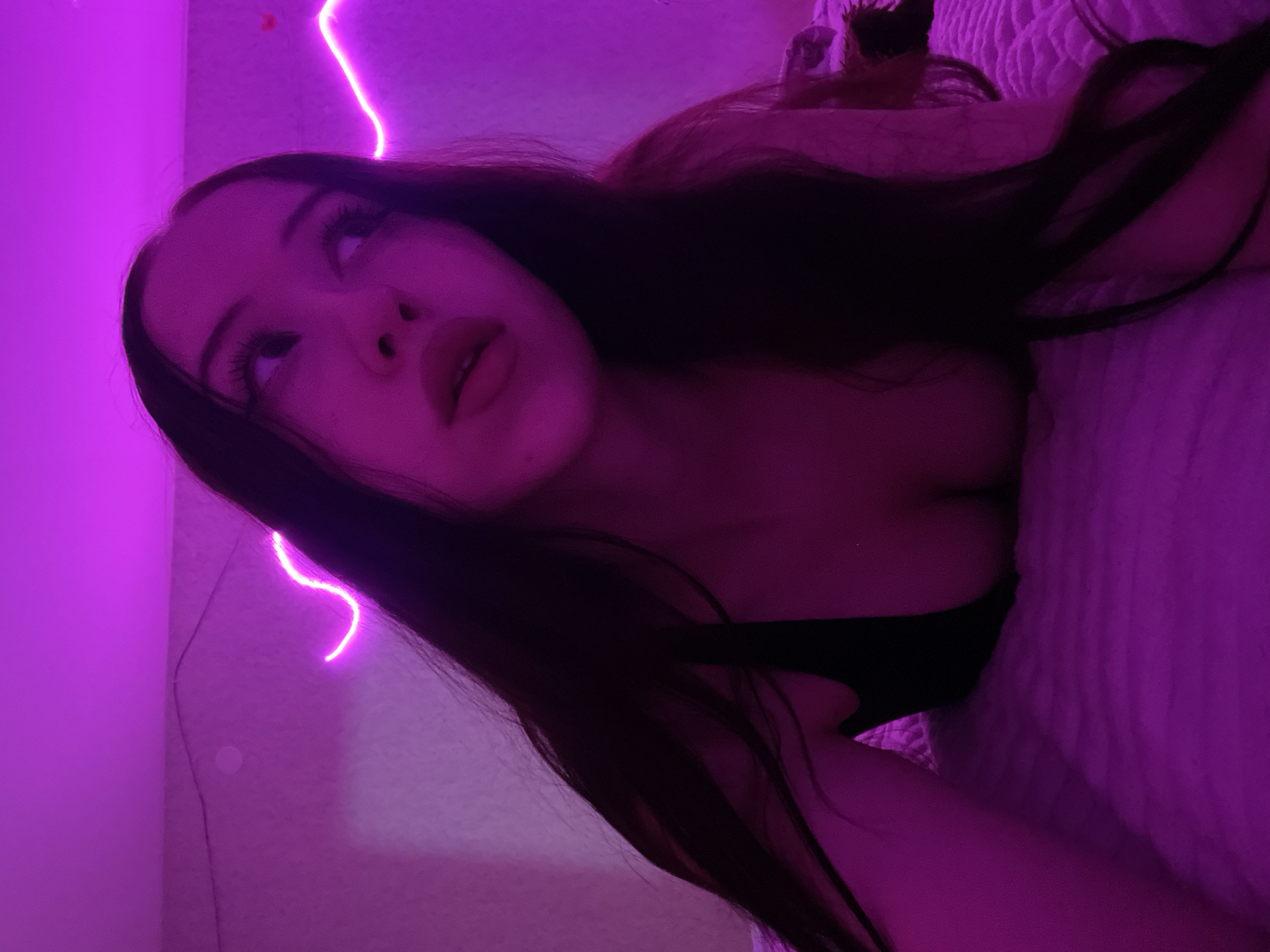 The image shows a young woman lying on a bed with pink neon lights illuminating the scene. Her dark hair is long and falls around her shoulders. She is looking upwards and to the side, with her mouth slightly open. The bedding appears to be textured and white. The background wall is also lit by the pink neon lights, which are shaped into a wavy line. There are no discernible elements in the image that would allow for an accurate determination of the location within Votkinsk, Russia.