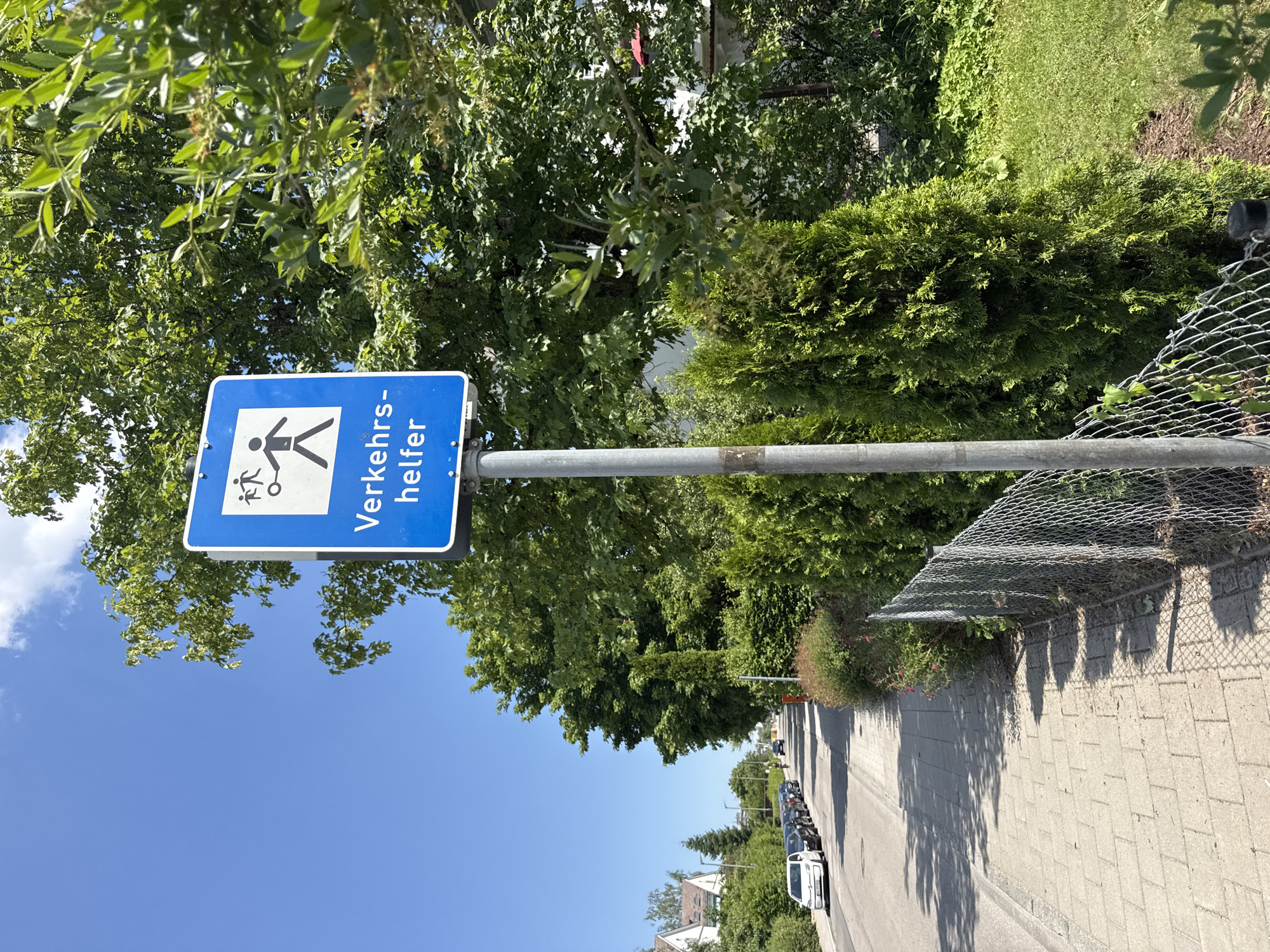 The image shows a street scene in Höhenkirchen-Siegertsbrunn, Germany. A blue sign with white text and symbols is prominently displayed on a metal pole in the foreground. The sign reads "Verkehrshelfer" and features two pictograms: one of two people walking with a child, and another of a person directing traffic. To the left of the sign is a paved sidewalk with a row of parked cars lining the adjacent street. The street appears to be a residential area, with houses and lush green trees visible in the background. A chain-link fence runs along the edge of the sidewalk, separating it from a well-maintained garden with manicured shrubs and trees. The sky is clear and blue, indicating a sunny day. The overall impression is of a quiet, suburban street on a bright summer day.