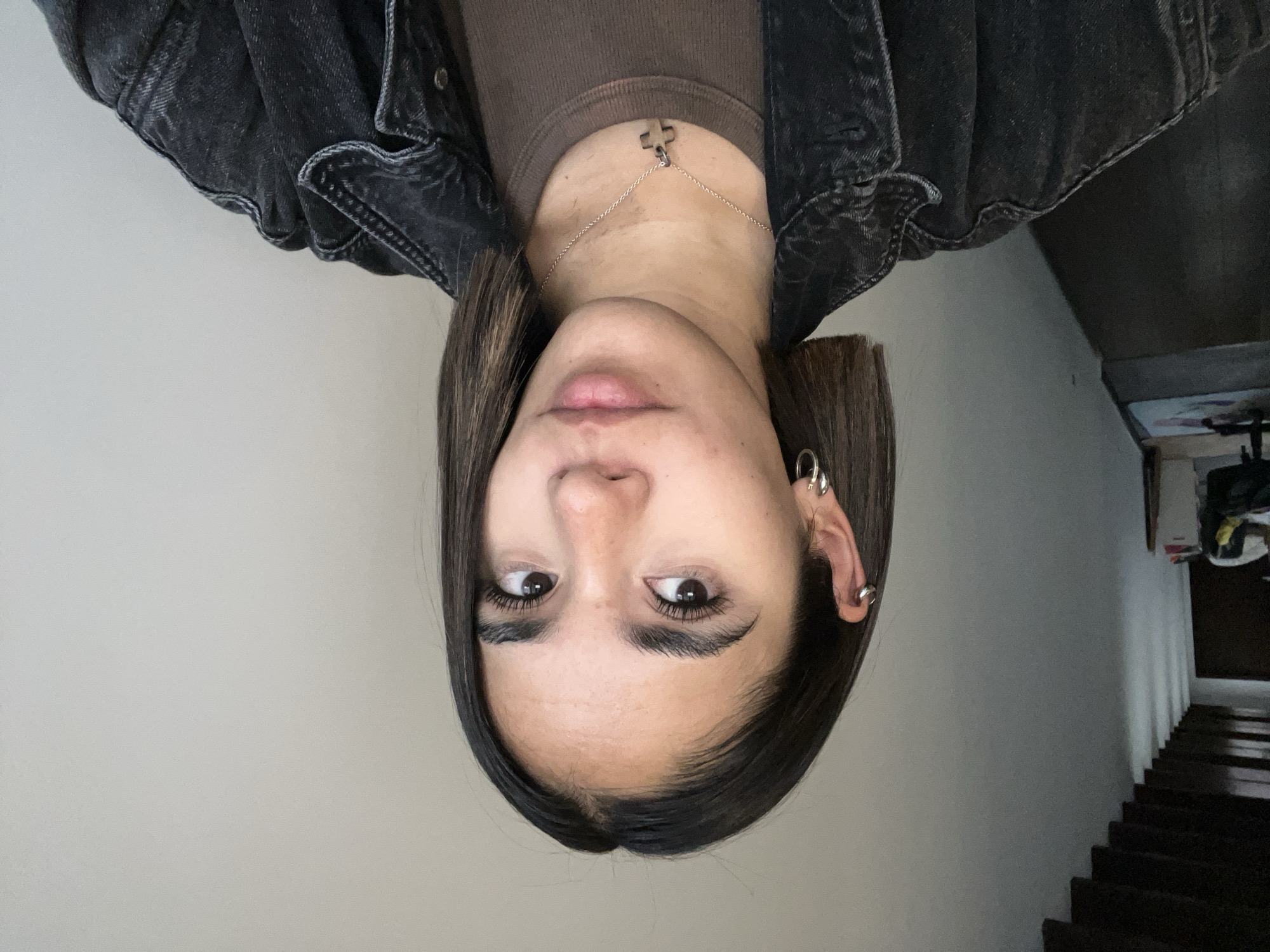 A person with straight, dark hair and light skin is facing the camera with a neutral expression. They are wearing a brown t-shirt and a dark denim jacket. A silver chain necklace with a cross pendant is visible. The person has multiple silver earrings on their left ear. The background is blurry, but a white wall and a staircase leading downward are discernible. The location is suggested as San Bernardino Tlaxcalancingo, Mexico, but there's no specific imagery to confirm this definitively.