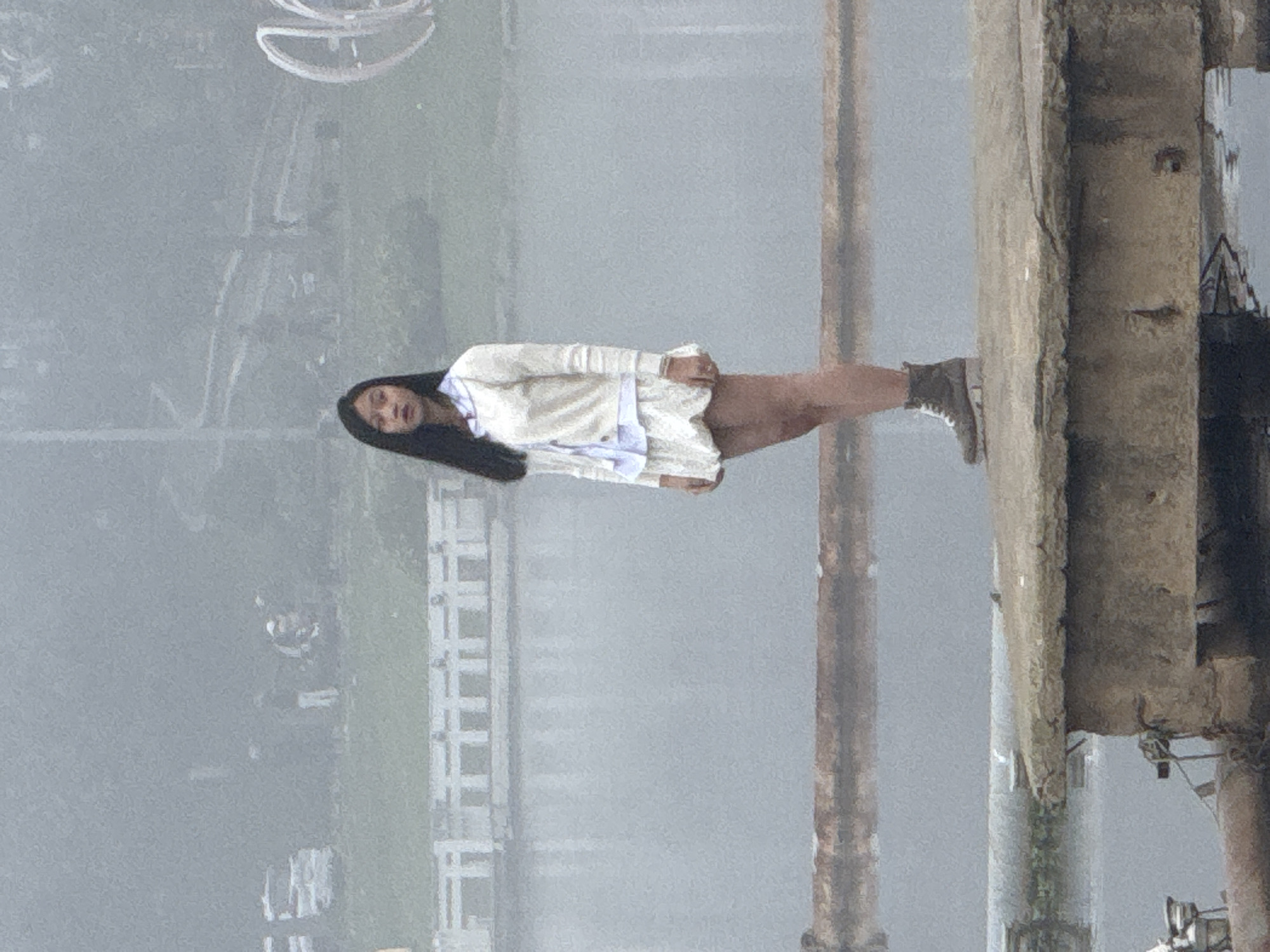 A young Asian woman with long, dark hair stands on a concrete platform at the edge of a body of water. She is wearing a cream-colored cardigan over a light blue collared shirt and a cream-colored skirt with a ruffled hem. Her footwear consists of dark, lace-up boots. The background is obscured by a dense fog, but a white railing and a distant, blurred cityscape are faintly visible. The water appears calm, reflecting the muted tones of the overcast sky. The platform itself is weathered and cracked, showing signs of age and exposure to the elements. There are large, rusty pipes visible in the foreground. The scene conveys a sense of quietness and isolation due to the fog and the woman's solitary presence.