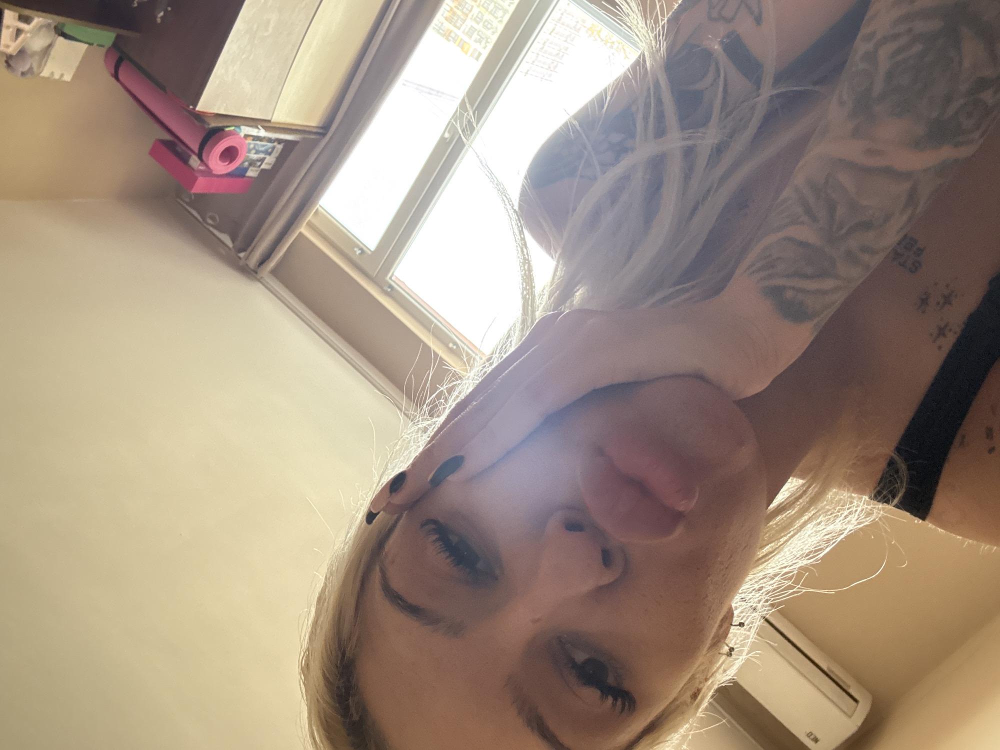 A close-up, high-angle selfie shows a fair-skinned person with blonde hair. The person's left hand is near their forehead, and their face is partially obscured. Tattoos are visible on their arm and shoulder. The person appears to be indoors, with a window visible in the background, letting in bright light. A rolled-up pink yoga mat is sitting on a stack of books or boxes, and an air conditioning unit is visible on the ceiling. The location is unknown.