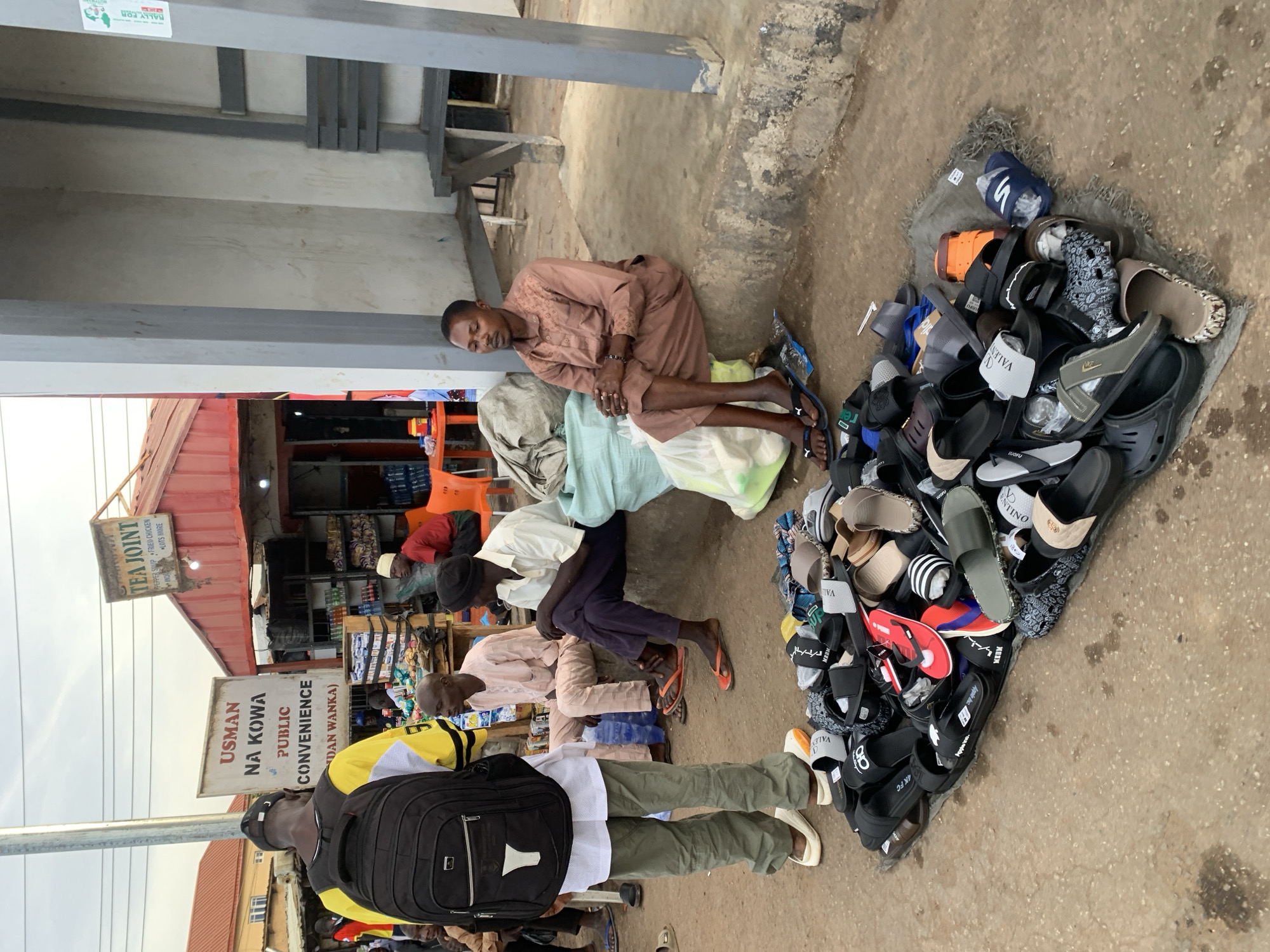 A street scene in Gura Top, Nigeria, shows several people in varying states of activity. A man sits on the ground next to a large display of sandals. Behind him, another man is sitting, and further back, a group of people is gathered near a building identified by signs as "USMAN NA KOWA PUBLIC CONVENIENCE" and "TEA JOINT". In the foreground, another man with a backpack walks past the sandal display.
