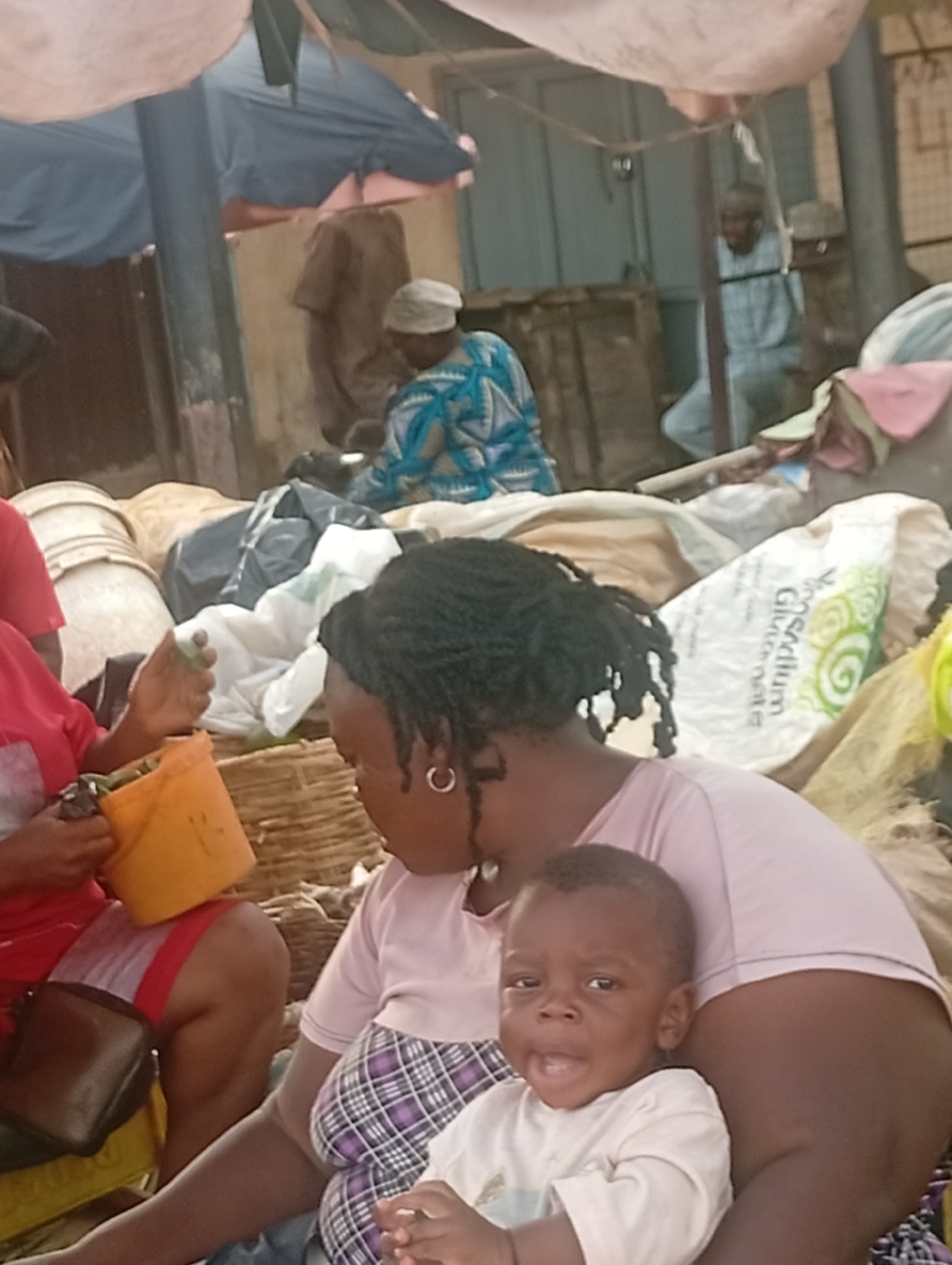 In Sabon Tasha, Nigeria, a woman with dreadlocks and hoop earrings sits, holding a baby on her lap. She is wearing a light purple shirt and a black and purple plaid skirt. The baby wears a light-colored shirt. The woman sits near a yellow bucket filled with green produce. Behind them, there is a man in a blue patterned garment and another man in a light blue outfit. Various sacks and containers are also visible. The scene appears to be outdoors under some kind of overhead covering or awning.