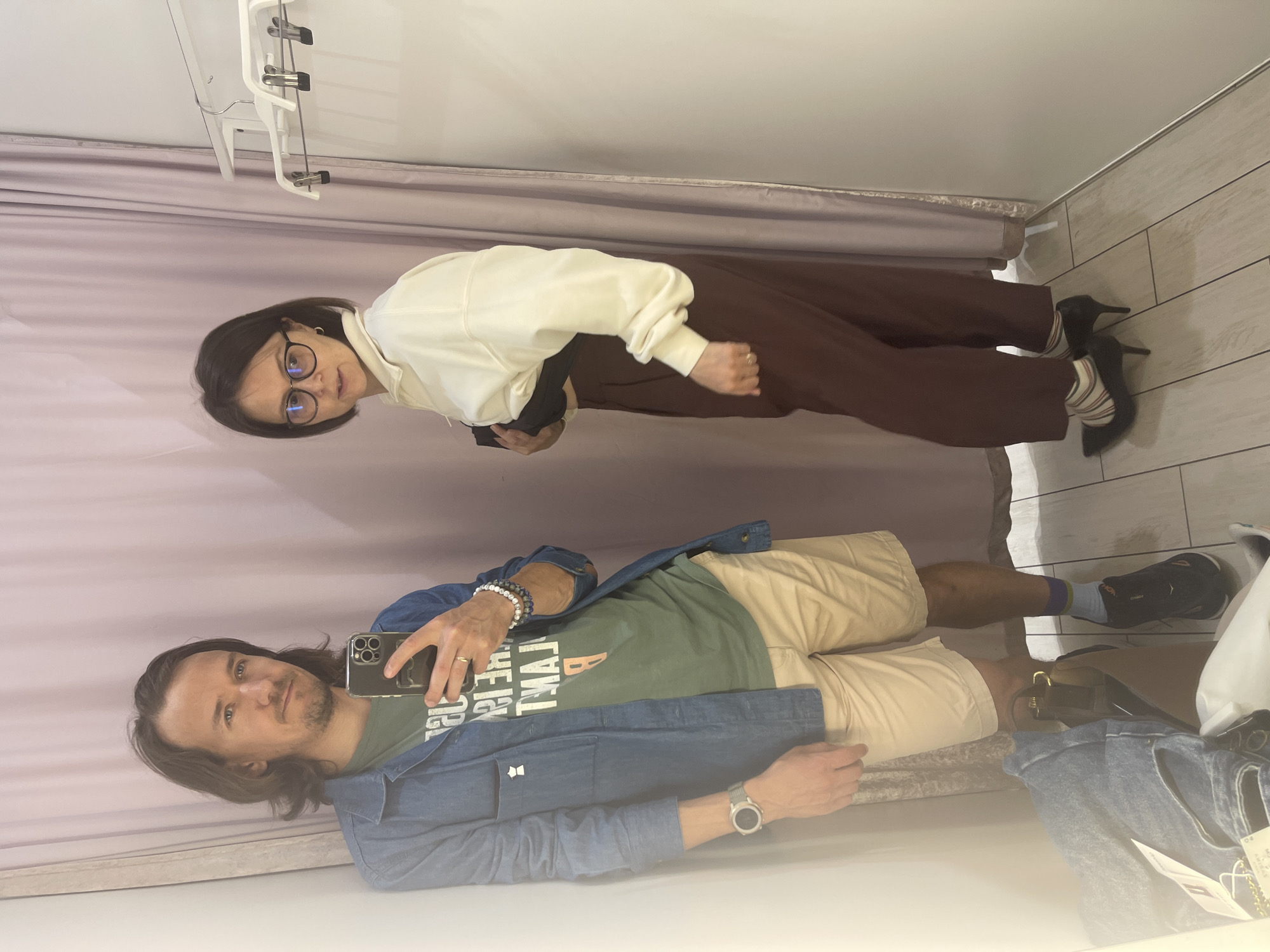 A man and a woman are captured in a mirror selfie in what appears to be a clothing store fitting room.
The man, holding the phone, has long hair and is wearing a green t-shirt, a blue denim jacket, and tan shorts. He also has a wristwatch on his left wrist.
The woman is wearing glasses, a white top, and brown trousers, paired with striped socks and black high heels.
The fitting room has a curtain and tile-like walls. Clothing hangers are visible above. The floor is off-white with what appear to be gray seams. There are clothes and shopping bags.
