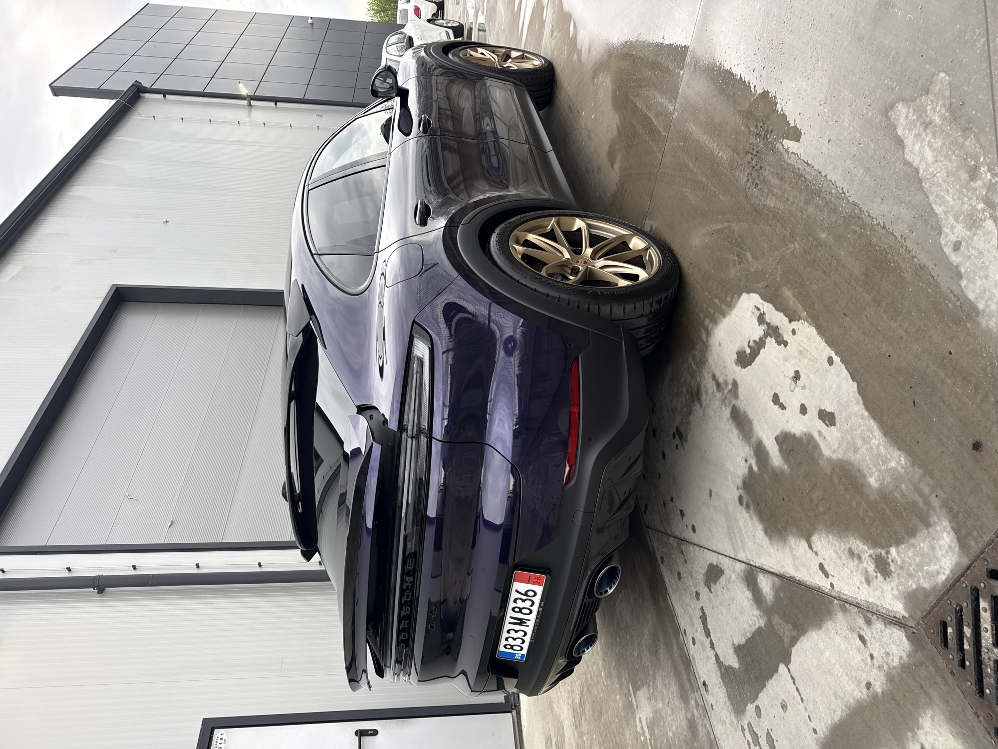 A dark purple Porsche Cayenne Turbo is parked on a wet concrete surface next to a corrugated metal building in Plovdiv, Bulgaria. The car's rear end is visible, showing the license plate "833 M 836" with the initials "BG" indicating Bulgaria, and a small "26" in the upper right corner. The car has gold-colored rims and quad exhaust pipes. Patches of water and tire tracks are visible on the ground. The building has a gray roof and a closed, large door with a smaller service door below. Another car, partially visible, is parked in the background. The lighting suggests it is an overcast day.