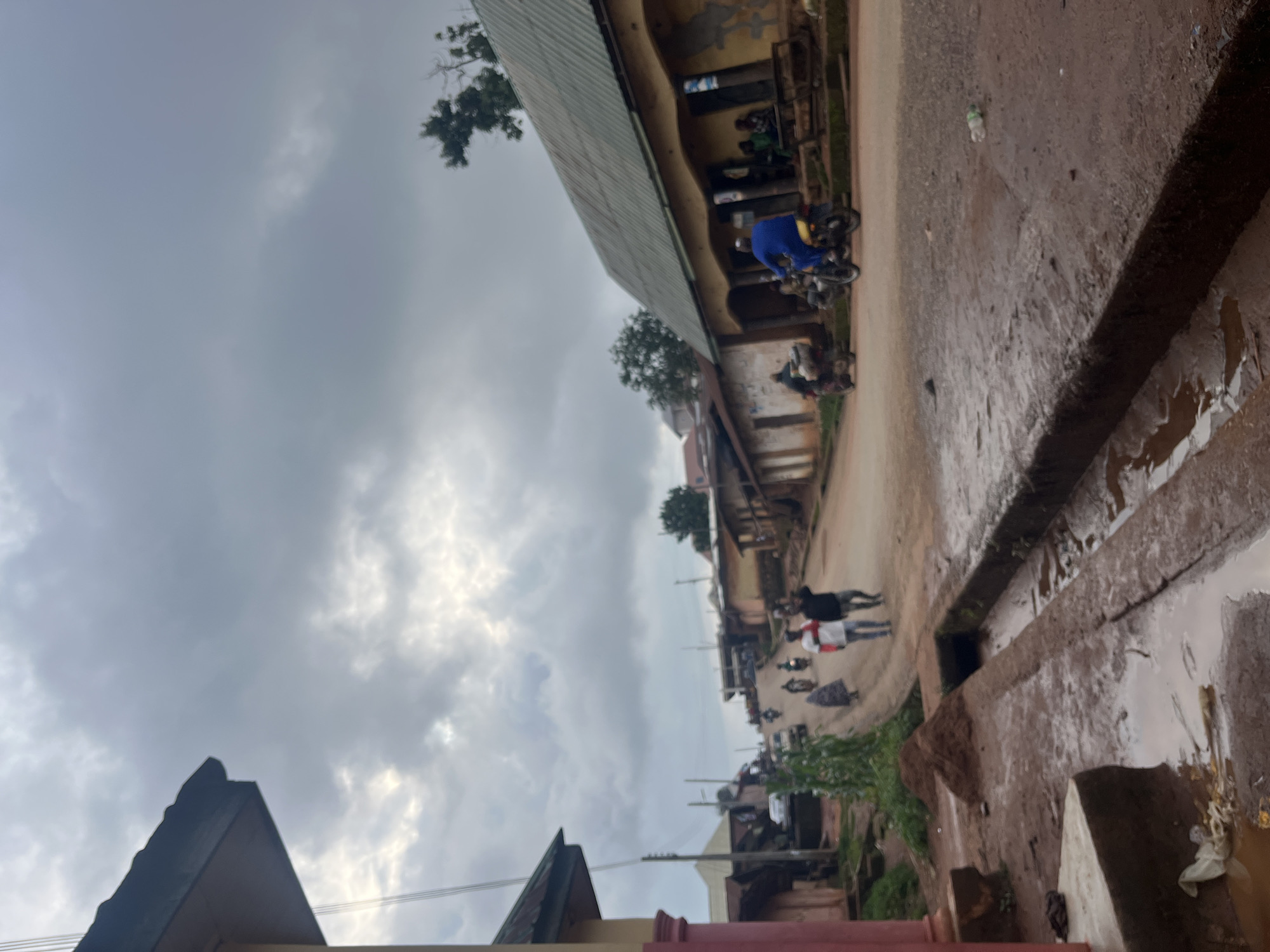 The image depicts a street scene in Gembu, Nigeria, under a cloudy sky suggesting either early morning or late afternoon. The street is unpaved and appears to be wet, with puddles indicating recent rain. Buildings line the street, with some showing signs of wear and tear.
Several people are visible, some walking and others on motorcycles. One person in a blue garment is on a motorcycle, while others are seen in groups or individually on the street. There are multiple motorcycles present, suggesting they are a common mode of transportation in this area. Greenery is visible in patches along the street, indicating some vegetation.
The overall atmosphere is one of everyday activity, with people going about their business despite the overcast weather. The lighting is subdued due to the clouds, and there is no clear indication of any specific event. The wet ground and puddles are a prominent detail, hinting at the tropical climate of the region. No visible text is discernible in the image. The composition of the photograph, with its tilted perspective, adds a dynamic feel to the scene.
