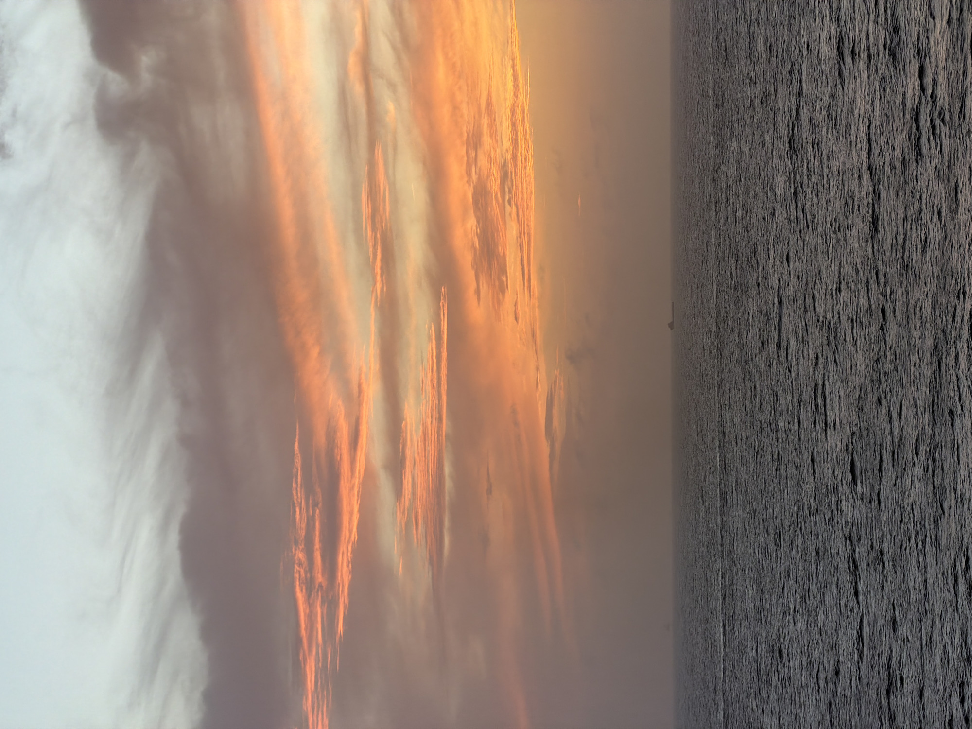 The image depicts a landscape with a striking contrast between the sky and the ground. The sky, taking up a significant portion of the frame, is filled with textured clouds illuminated by a warm, orange light, possibly indicating sunrise or sunset. The clouds are layered and streaked, creating a dynamic visual effect. On the right side of the image, the ground is characterized by a dark, rough surface. The texture appears to be densely packed and uneven, possibly representing barren land or a rocky terrain. The scene is located in the city of Unknown, Unknown.