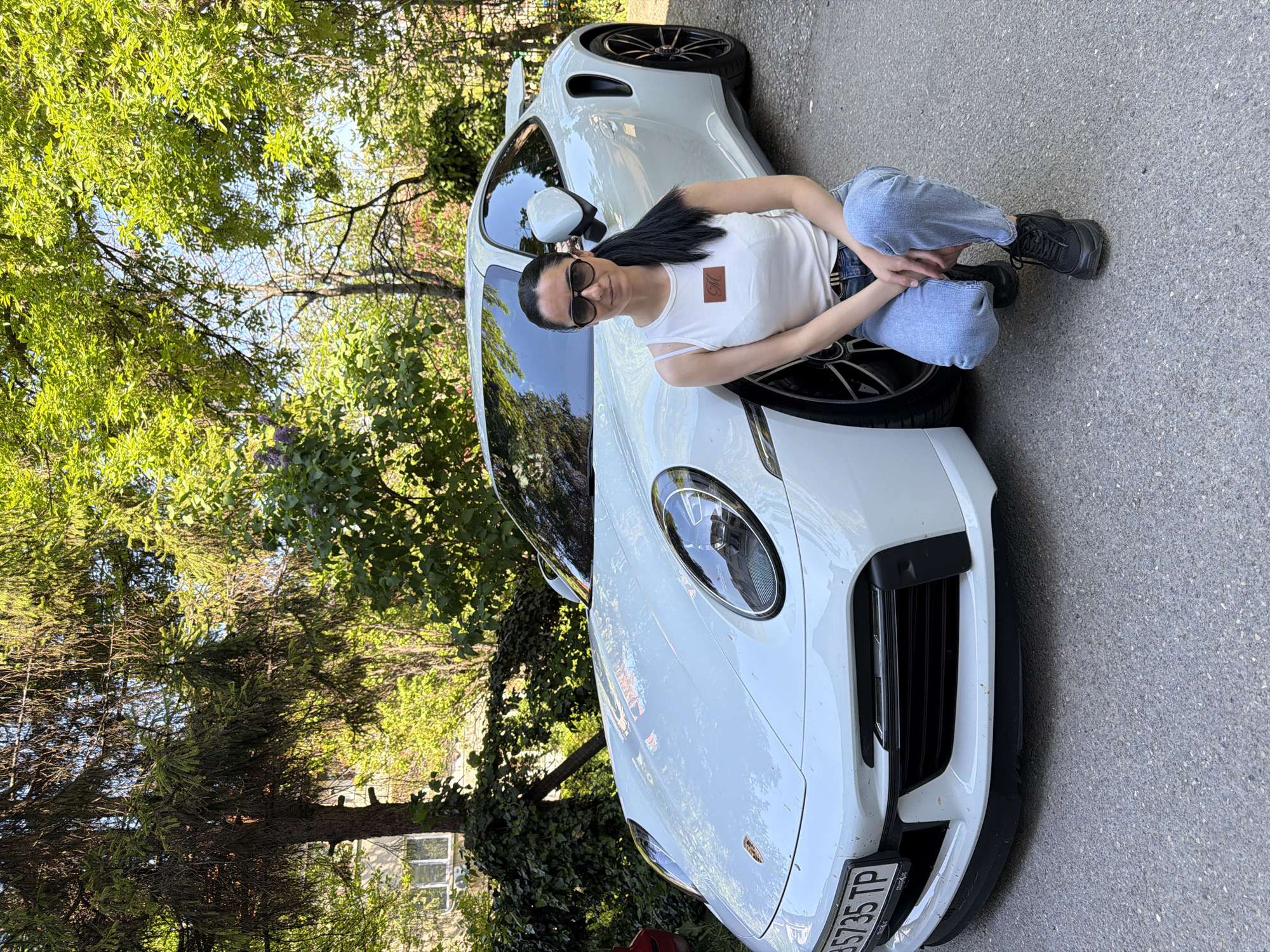 A woman is crouched beside a white Porsche 911 Turbo S. She is wearing jeans, a white tank top with a logo, and sunglasses. The car is parked on a paved street near trees. The license plate of the car reads “PB5735 TP”. The location is Pleven, Bulgaria.