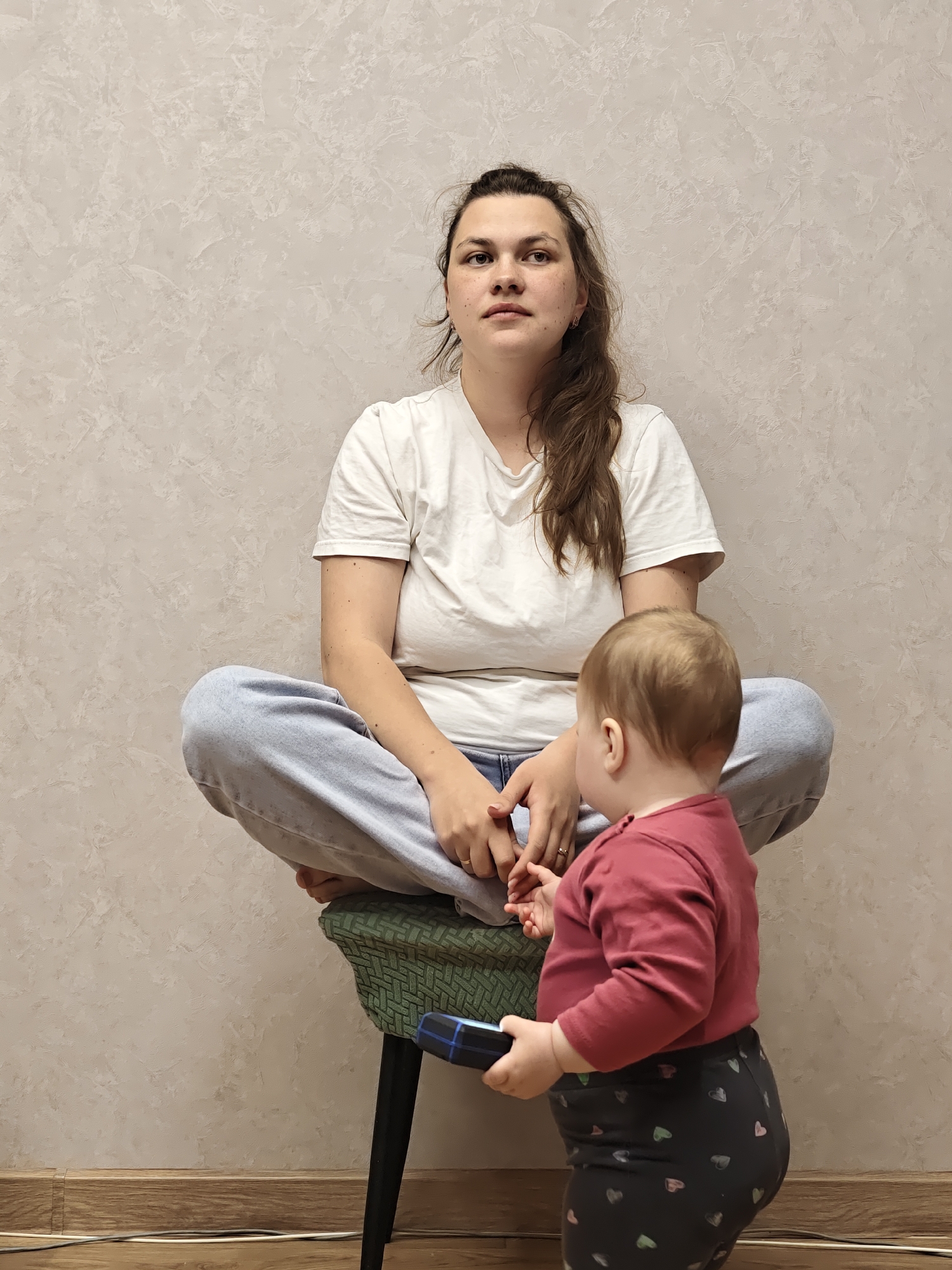 An indoor scene in Chaikovsky, Russia, features an adult woman and a baby in a domestic setting. The woman is seated cross-legged on a dark green patterned stool, facing forward with a neutral, slightly contemplative gaze directed upwards and to her right. She has long, light brown hair, partially tied back, and wears a simple white short-sleeved t-shirt and light blue jeans.
In front of her, a baby stands, facing away from the viewer and towards the woman. The baby is dressed in a dark red long-sleeved shirt and dark leggings patterned with numerous small, light-colored hearts. It holds a small, dark blue object, possibly a toy or remote control, in its hands.
The background consists of a light beige wall with a subtly textured finish, resembling decorative plaster. The floor is made of light-colored wood, bordered by a matching baseboard. A few dark electrical cables are visible resting on the floor near the wall. The lighting suggests daytime. The overall atmosphere is calm and domestic, capturing a quiet moment between the two. There is no visible text.