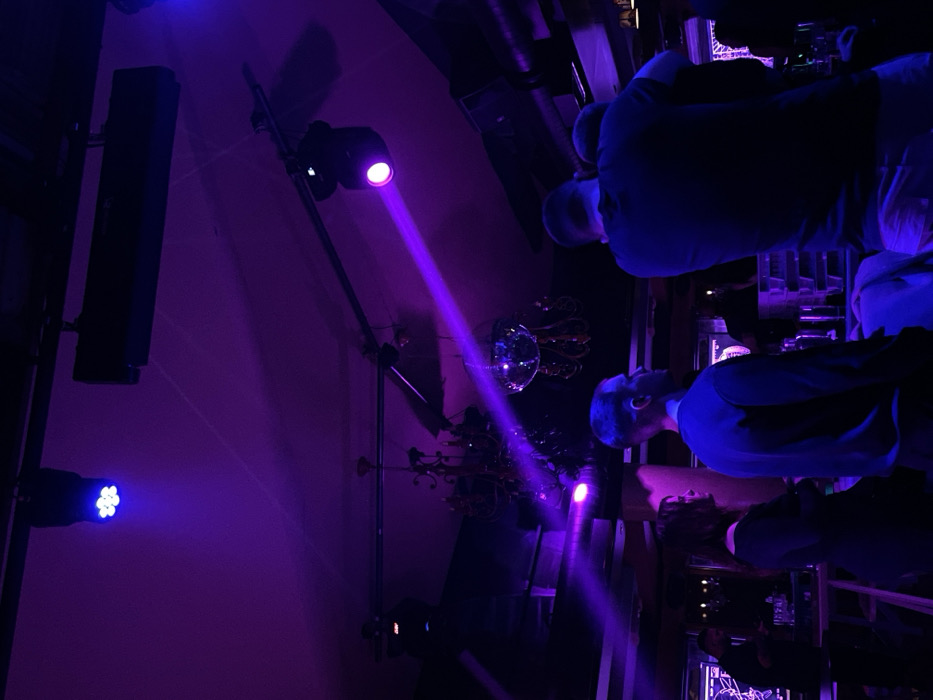 This media file depicts an indoor social gathering, likely at night, in Drapetsona, Greece. The scene is illuminated by vibrant artificial lighting, predominantly in shades of purple and blue, creating a dynamic and low-light atmosphere.
On the left side of the frame, a tall, black rectangular speaker is mounted on a stand. Nearby, several spotlights are visible. One spotlight positioned centrally emits a strong, distinct purple beam that cuts diagonally across the room, while another closer to the foreground projects a softer blue light. These lighting fixtures and the speaker are supported by dark stands or a truss system.
In the mid-ground, a small, reflective disco ball hangs, catching and scattering light, with some dark foliage or decorative elements visible beneath it. On the right side of the image, several individuals are present, seen mostly from the back or side. They appear to be standing or leaning at what looks like a bar or counter, which is dimly lit and shows hints of bottles or equipment. The people are dressed in dark attire, and their faces are largely obscured by shadows and the colored lights. The overall impression is of an active event taking place in an entertainment venue. No visible text is present in the image.