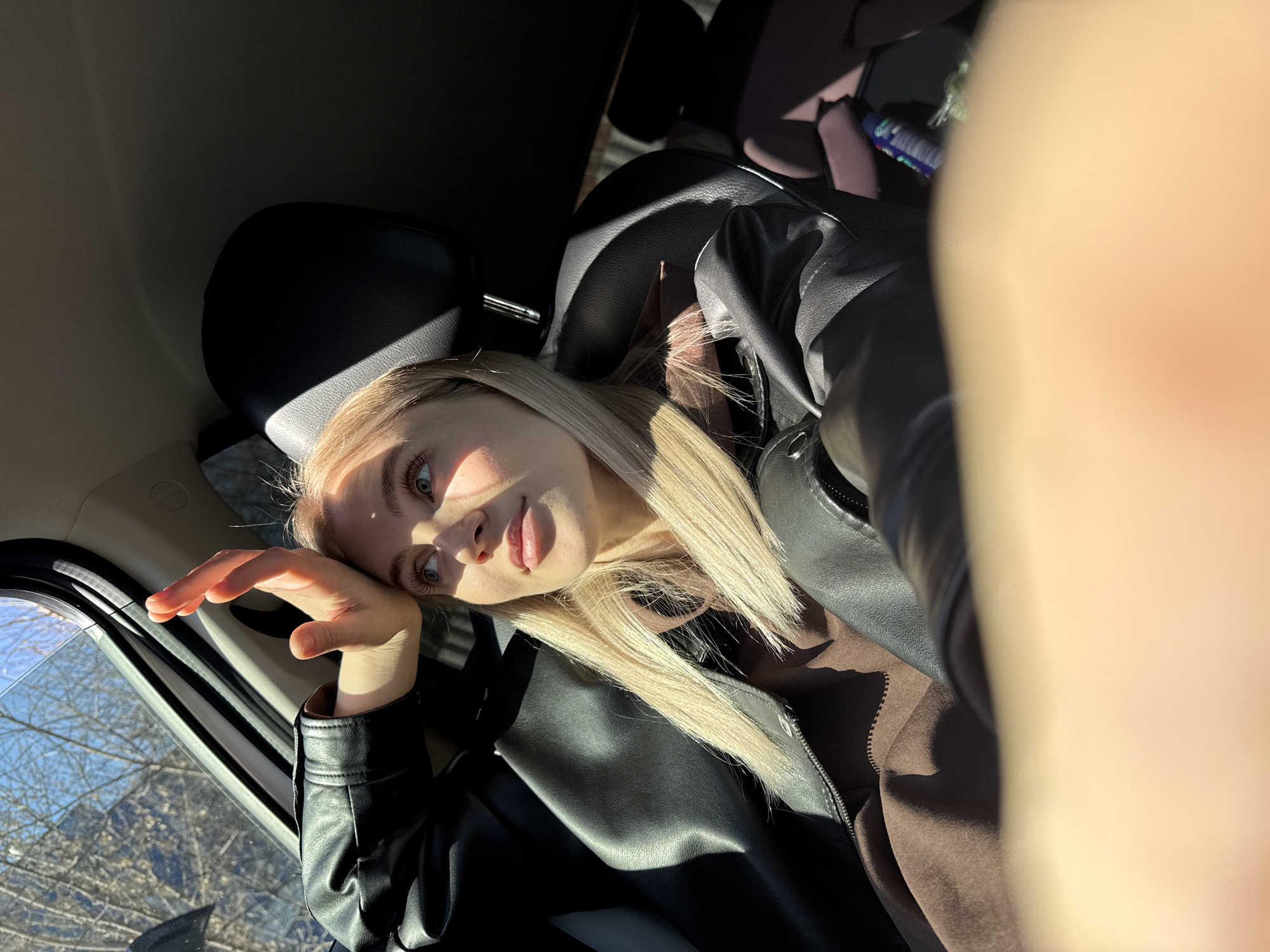 A fair-skinned young woman with blonde hair is in the back seat of a vehicle. She is wearing a black leather jacket over a brown hooded sweatshirt. Her left hand is near the window, which shows bare trees and a blue sky. A blurry object is in the foreground to the right, suggesting the photo was taken with a phone. Based on the interior of the vehicle, it is possibly a modern car. The location in Izhevsk, Russia is not discernible from this image alone. The interaction and action are a selfie taken inside the vehicle. There are no additional people present.