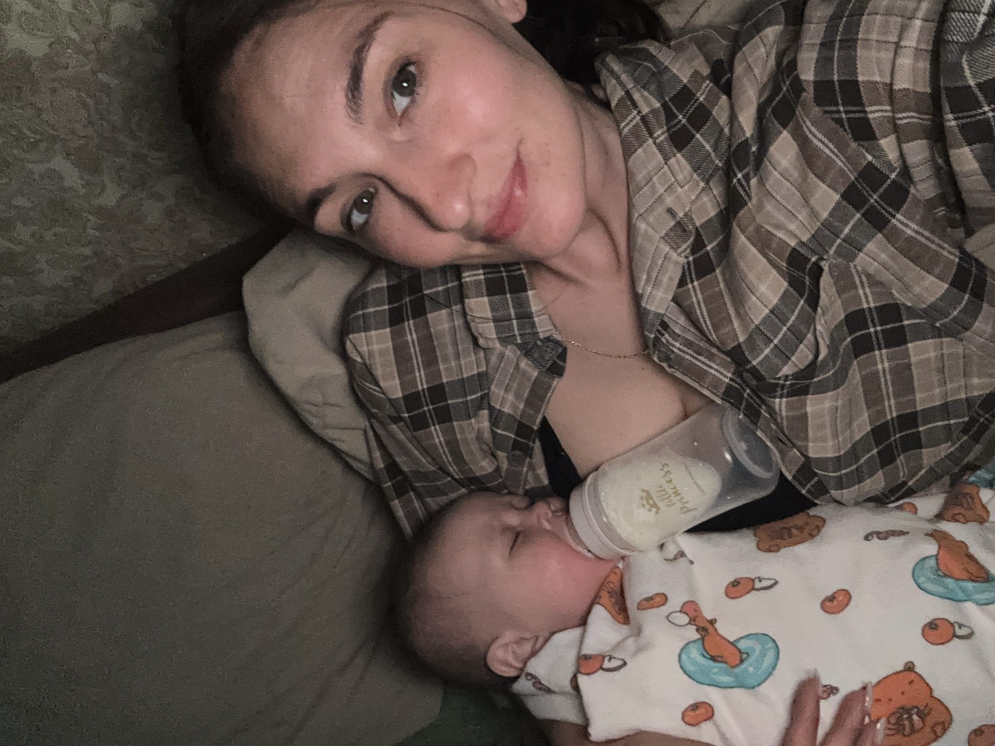 A woman and a baby are indoors. The woman lies on her side, angled towards the camera. She has light skin, brown eyes, and brown hair pulled back. She is wearing a plaid shirt. A baby lies next to her, bottle feeding. The baby is wearing a white onesie. The environment appears to be a bed or soft surface. There is no visible evidence to confirm this image being taken in Sochi, Russia.