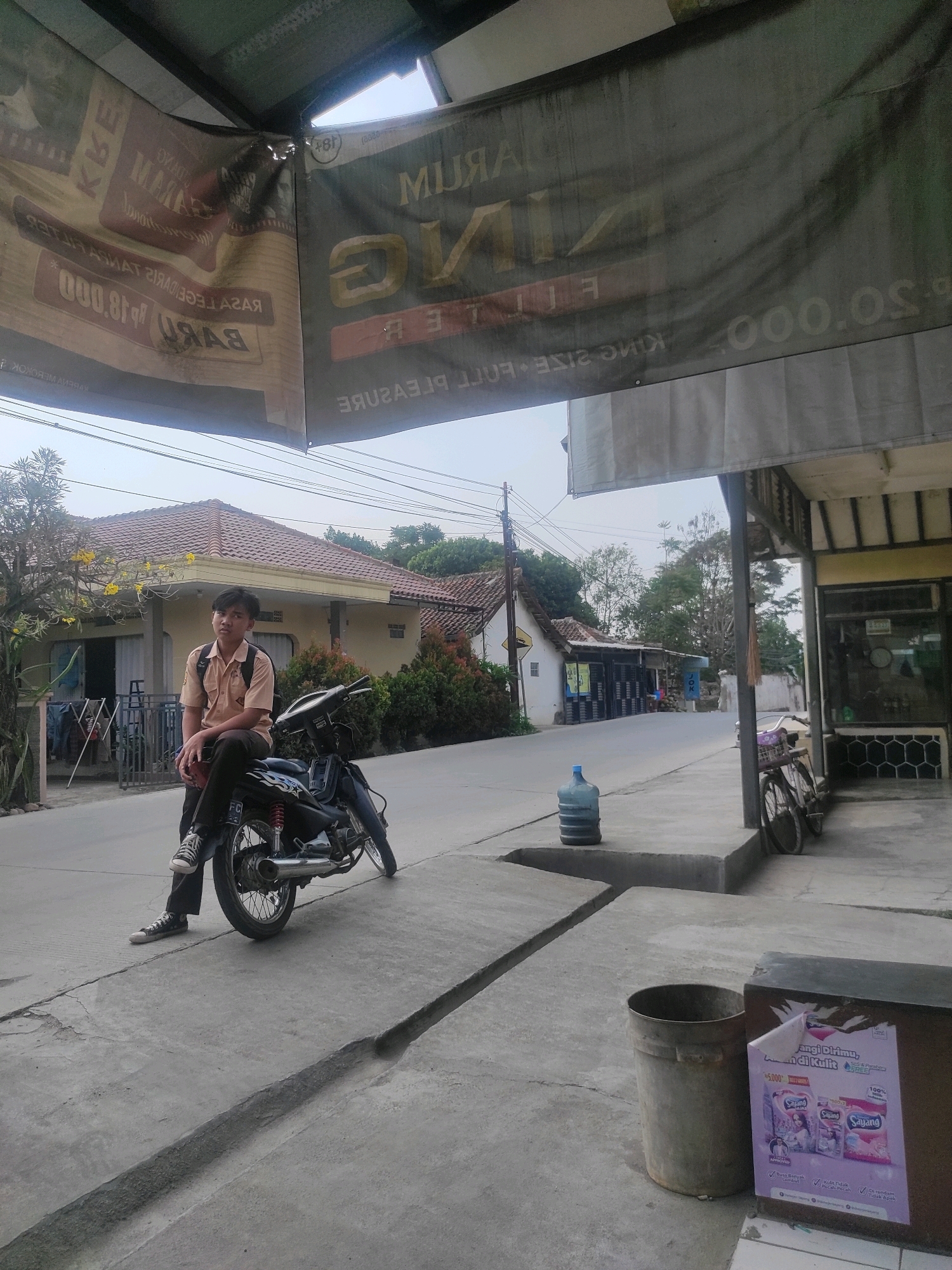 An outdoor shot captures a young Indonesian male in a school uniform, consisting of a light brown short-sleeved shirt and dark pants, sitting on a black motorcycle. He is positioned on the sidewalk next to a street in Ngamprah, Indonesia, with a backpack on his shoulders. The motorcycle is parked on a concrete surface with a drainage ditch in front of it.
Behind the boy, a modest yellow house with a red-tiled roof is visible. Across the street, a row of shops with dark storefronts and a wider street with utility poles and overhead wires suggest a moderately busy commercial area. A large blue water cooler bottle stands near the sidewalk.
Above the scene, a large banner hangs, obscuring some of the sky. The banner features text and graphics, but much of it is illegible due to its condition and the angle of the photograph. Another banner, partially visible on the left, displays text related to "RAJA" and "000.80.000". A bicycle is parked near one of the shops. The overall lighting suggests it is daytime, possibly late afternoon, given the soft light and shadows.