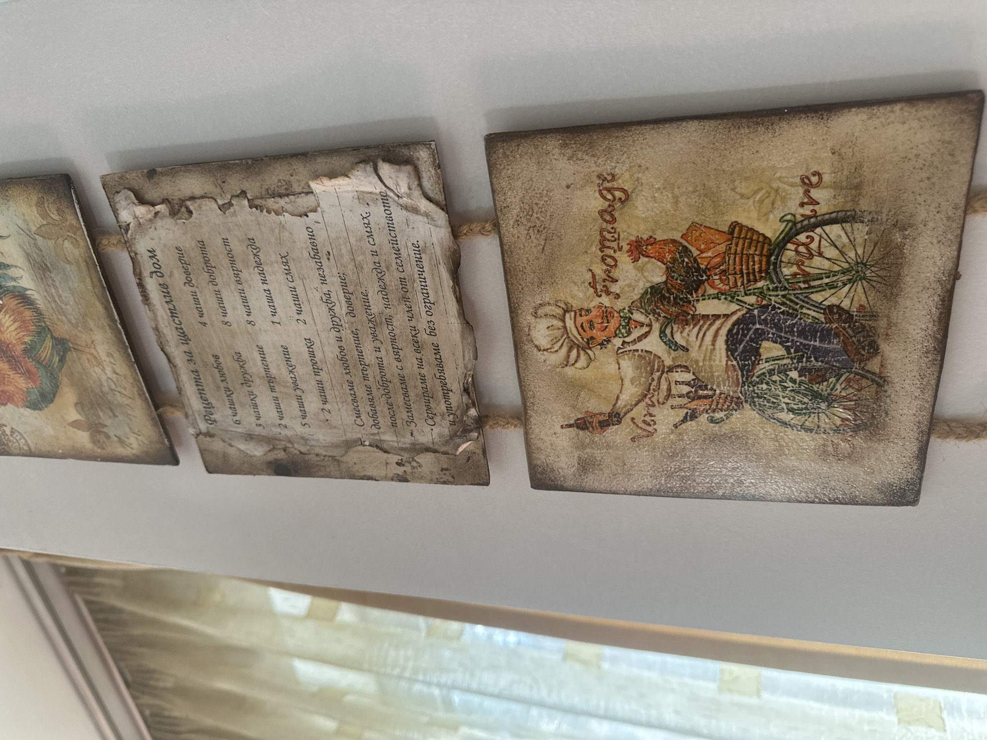 The media file captures three rustic, rectangular decorative plaques connected by a braided jute string, displayed against a plain, light-colored background in an indoor setting in Sofia, Bulgaria. The image is taken from a slightly downward-angled perspective.
The plaques have a vintage, distressed appearance, mimicking aged paper with torn edges and faded textures, consistent with a decoupage or antique craft style.
The central plaque features text in Bulgarian, titled "Рецепта за щастлив дом" (Recipe for a happy home). It lists metaphorical ingredients for a joyful household, including "4 чаши доверие" (4 cups of trust), "8 чаши доброта" (8 cups of kindness), "6 чашки любов" (6 cups of love), "2 чаши прошка" (2 cups of forgiveness), and "2 чаши смях" (2 cups of laughter). The text continues with instructions: "Смесваме любов и дружба, незабавно добавяме търпение, доверие; -после доброта и уважение.. Замесваме с вярност, надежда и смях. Сервираме на всеки член от семейството и употребяваме без ограничение." (Mix love and friendship, immediately add patience, trust; then kindness and respect... Knead with faith, hope, and laughter. Serve to every family member and consume without restriction.)
The rightmost plaque displays an illustration with a culinary theme, featuring a character in a chef's hat, a bicycle, a basket, and a rooster. The prominent text on this plaque is "Fromage" (French for Cheese) in an elegant script, with a partial word "dire" also visible. A third plaque is partially visible on the far left. The decorative items evoke themes of home, family, and culinary enjoyment. A blurry reflection of an indoor light source or window is visible at the bottom edge of the frame.