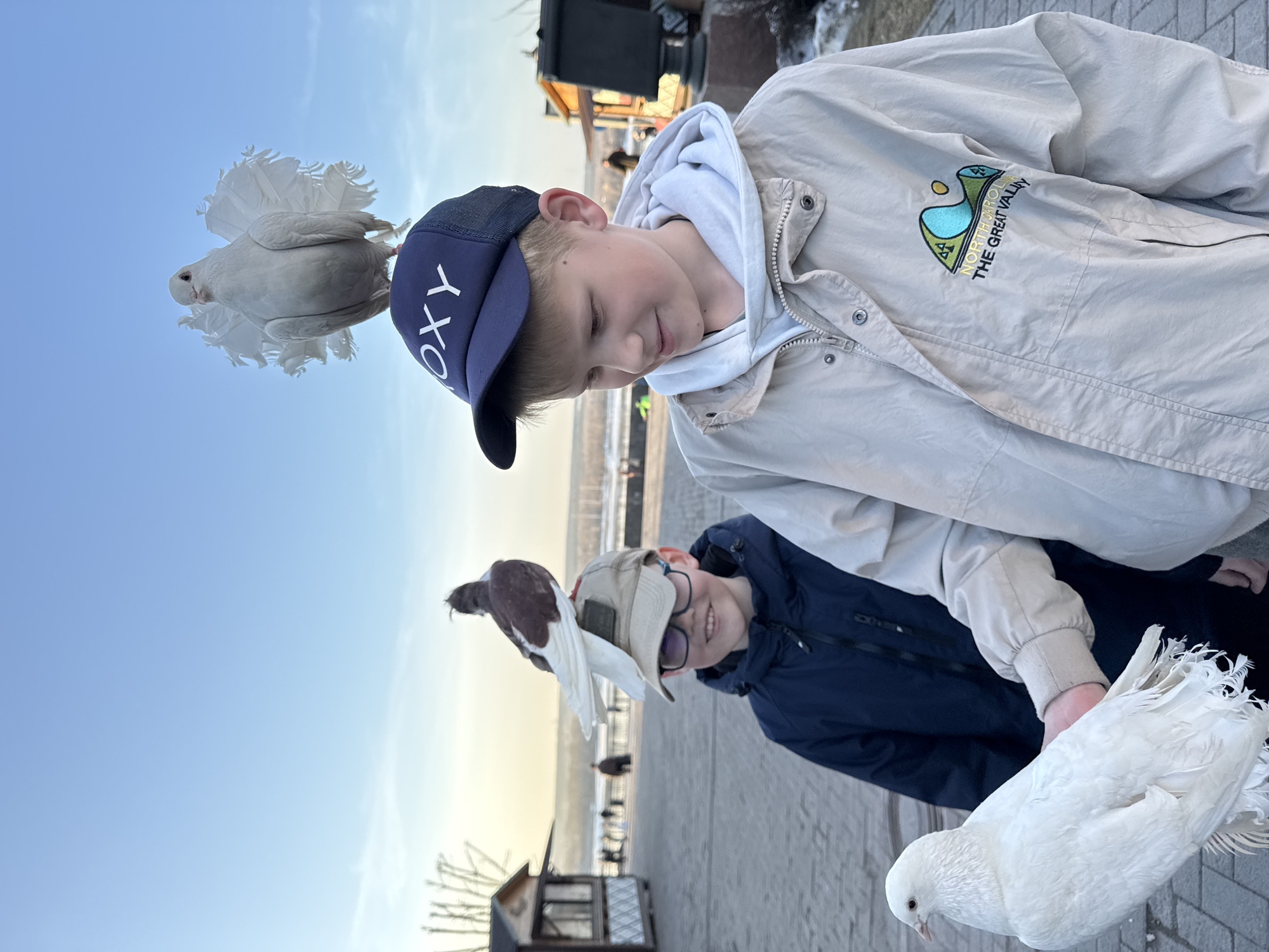 Two boys stand outside, interacting with pigeons. One boy wears a navy blue baseball cap with "ROXY" on it and a light-colored jacket over a white hoodie, looking downward with a slight smile. On his head sits a white pigeon. The other boy, wearing glasses and a tan baseball cap, smiles towards the viewer; he has a brown and white pigeon on his head. A white pigeon is in his hand. The background includes a paved area, a view of a body of water, and indistinct buildings or structures. It is the waterfront of the city of Perm, Russia.