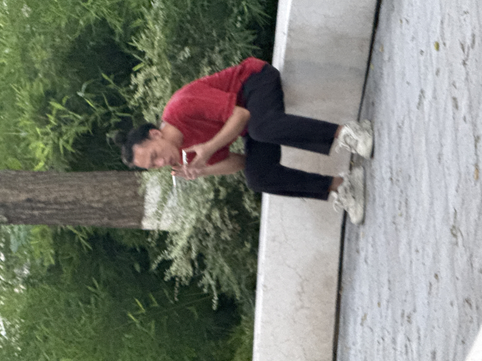 A person with dark hair pulled back in a bun is sitting on a low concrete wall, facing to the left and smoking a cigarette. They are wearing a red short-sleeved shirt and dark pants, with white athletic shoes. Behind them, a tree trunk with white paint around its base is visible, and beyond that, dense green foliage. The ground in front of the wall is a grey paved surface with scattered small debris. The scene appears to be an outdoor public space, possibly a park or plaza, in Yuzhong District, China.
