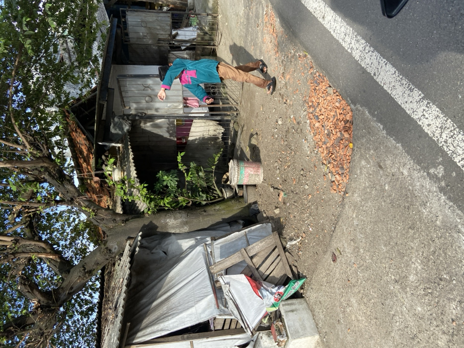 The scene is an alleyway in Sukatani, Indonesia, located at the coordinates -6.639 latitude and 107.408 longitude. A young man wearing a blue shirt, pink jacket, and black pants is walking away from the camera, exiting the alleyway and onto the paved street. He has his left arm raised and is gesturing with his hand. The alleyway is lined on both sides with small, simple structures constructed of brick and corrugated metal. A large tree grows in the center of the alleyway and casts a shadow across the ground. A pile of broken bricks lies near the edge of the paved road. The ground in the alley is covered with dirt, debris, and fallen leaves. The scene is sparsely populated with no other people visible. The street beyond the alleyway is paved and marked with a white line down the center. The sky is bright and clear. The sun is shining. The image has a slightly obscured view of a vehicle in the background.