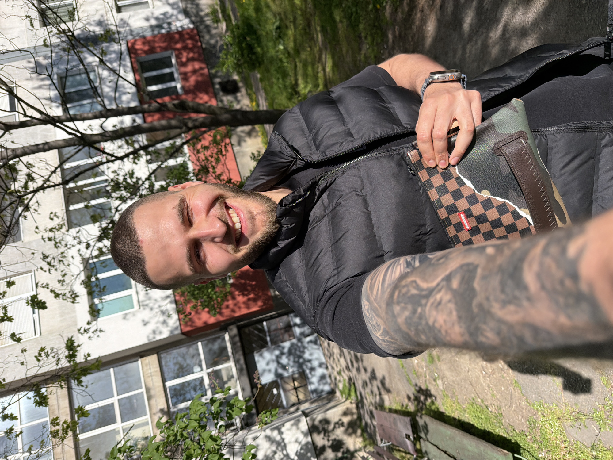 A young man with a short beard and closely-cropped dark hair smiles broadly at the camera, capturing a selfie outdoors in Sofia, Bulgaria. He wears a black puffer vest over a black t-shirt. His left arm, prominently tattooed with a full sleeve of dark, intricate designs, stretches into the foreground, and he wears a digital watch with an orange accent on his wrist. In his right hand, he holds a distinctive bag or pouch. This accessory features a split design: one section displays a brown and black checkerboard pattern with a small red "SPRAYGROUND" tag, while the other section is a camouflage pattern, separated by a jagged, torn-paper-like edge.
Behind the man, a multi-story building, likely an apartment block common in urban settings, stands with light-colored walls and numerous windows. A section of the building has a contrasting red facade. Bare tree branches are visible in the midground and foreground, partially obscuring the architecture. Green grass and foliage are present on the ground level, and a partial view of a bench can be seen. The bright sunlight indicates a clear and sunny day, casting strong shadows and highlighting the man's cheerful expression.