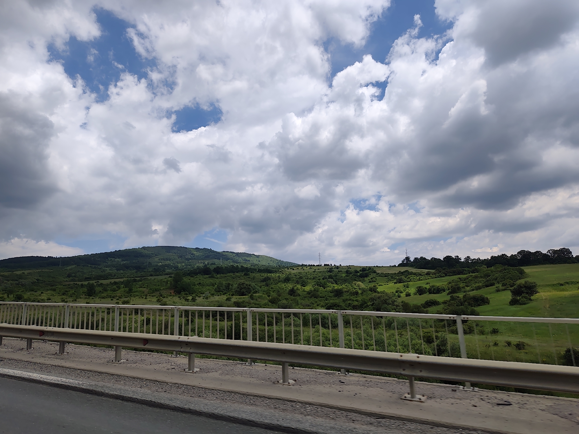 The image depicts a wide, sweeping landscape view from what appears to be a bridge or overpass. In the foreground, a metal guardrail runs horizontally across the frame, supported by vertical posts. The guardrail is a light metallic color and shows signs of wear. Below the guardrail is a concrete structure, and then the edge of a paved road.
The midground reveals a rolling, verdant landscape characterized by lush green hills dotted with trees and patches of open grassy fields. The terrain is uneven, with some areas appearing more densely wooded than others. In the distance, a larger, more prominent hill forms the backdrop, also covered in trees and vegetation. Power lines or transmission towers are visible on the horizon, suggesting the presence of infrastructure.
The sky above is a mix of bright blue patches and voluminous, heavy grey and white clouds. The clouds are dynamic, with varying degrees of thickness and texture, indicating a possibly changeable weather pattern. The overall impression is a rural, possibly somewhat undeveloped area under a dramatic sky. The location is identified as Dragichevo, Bulgaria, a village situated in the western part of the country, near Sofia.
