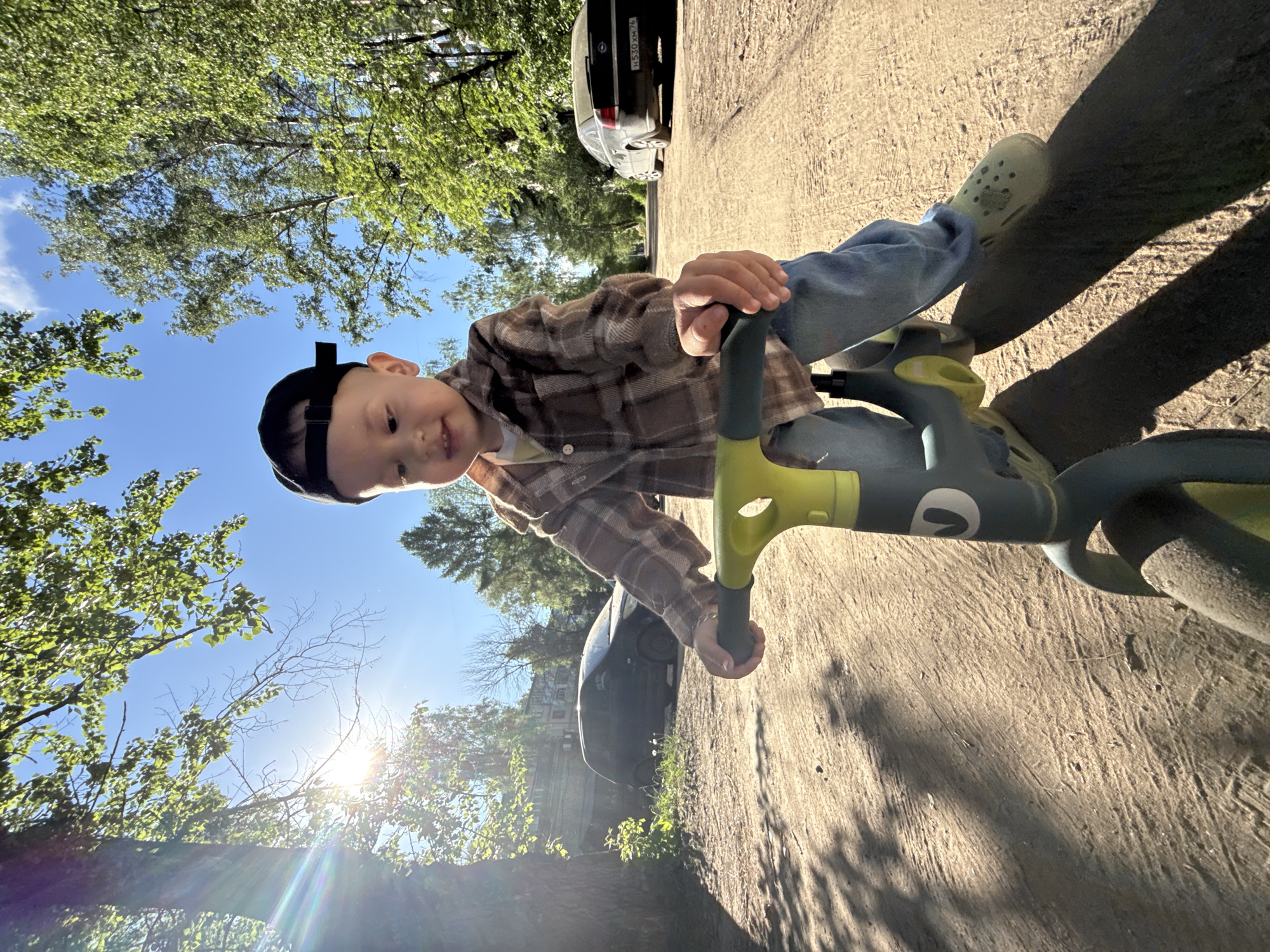 A young boy is riding a green balance bike on a paved road. He is wearing a brown plaid shirt, blue jeans, and a black cap. He has green Crocs on his feet. The road is in the city of Rusanovo, Russia, as evidenced by the license plate "H530 KM76" visible on a parked black car in the background, which indicates the Yaroslavl Oblast region of Russia. Trees line the road, and the sun is shining brightly.