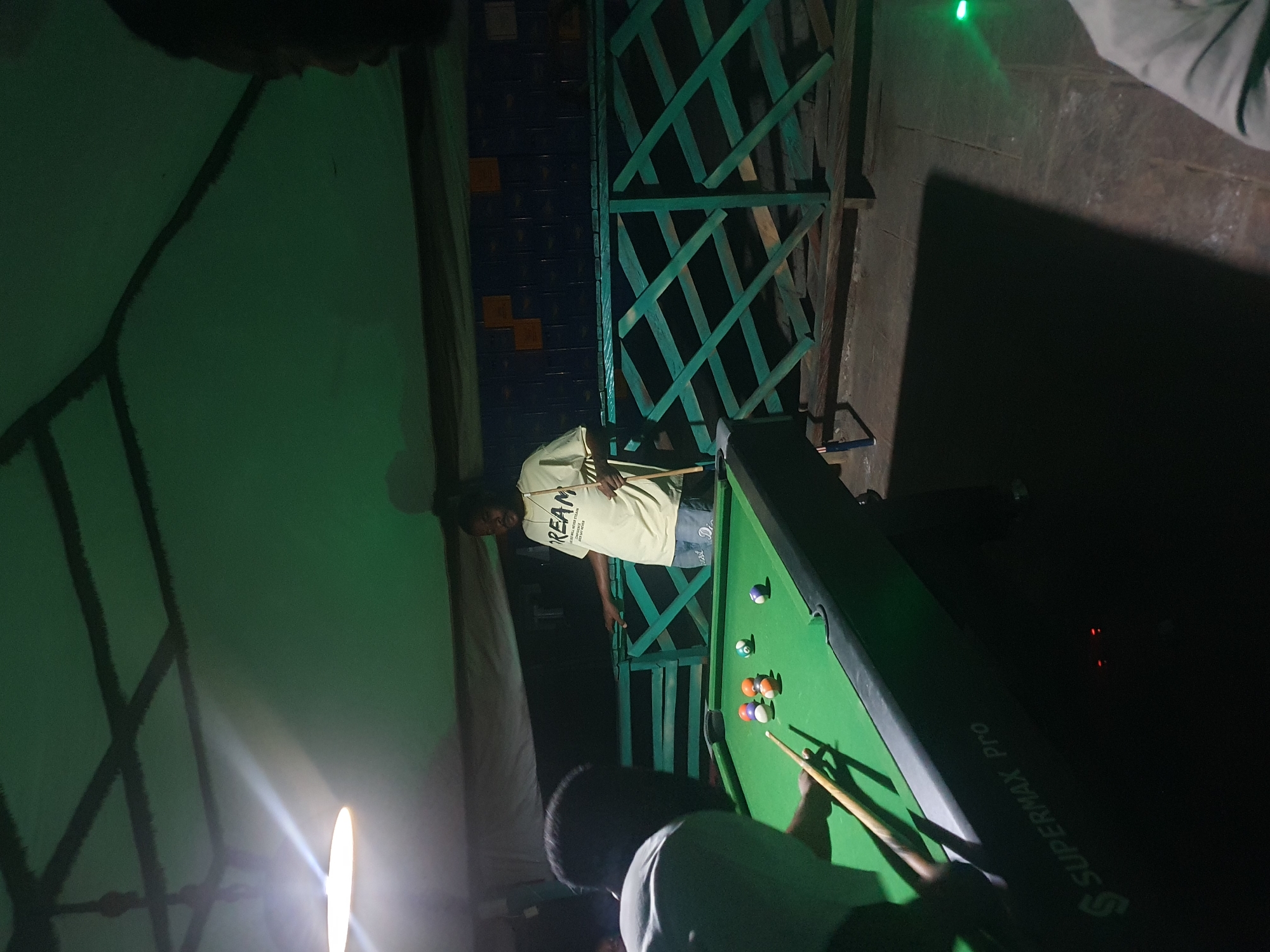 In Lokoja, Nigeria, a nighttime scene unfolds at a dimly lit, open-air establishment with a green tarp ceiling supported by dark beams. The focal point is a green pool table, branded "SUPERMAX Pro," where a game is in progress. Two individuals are engaged, with one person, partially visible from the back and wearing a light-colored shirt, holding a cue stick and poised to strike a ball. The other player, a woman wearing a yellow t-shirt with the word "DREAM" printed in black and denim shorts, stands across the table, also holding a cue. A collection of pool balls, including a red, an orange, a white with orange stripes, and a solid blue ball, are scattered on the felt. The background reveals a green lattice fence and a dark wall adorned with orange squares. A bright overhead light fixture casts a strong illumination on the left side of the scene, creating dramatic shadows. The overall ambiance suggests a casual gathering for a game of billiards.