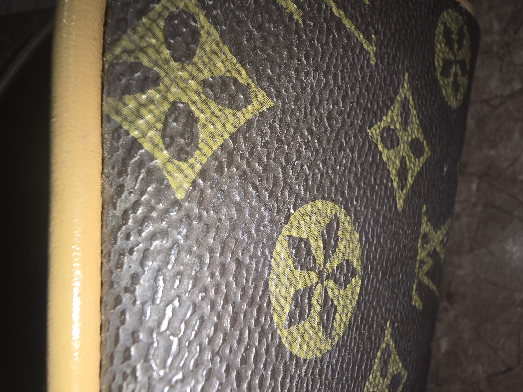 This is a close-up, high-angle, macro shot of a textured brown surface with a repeating yellow pattern and a tan-colored edge. The brown surface appears to be made of leather or a similar material with a dimpled texture. The yellow pattern consists of diamond shapes with circular elements inside, and also circular shapes with floral-like designs. The tan edge runs horizontally across the top of the frame. The lighting is harsh, creating bright highlights on the textured surface and the tan edge. The image does not provide any information about the location in the city of Ulan-Ude, Russia, as it is an isolated detail of an object. There are no people, actions, or interactions visible.