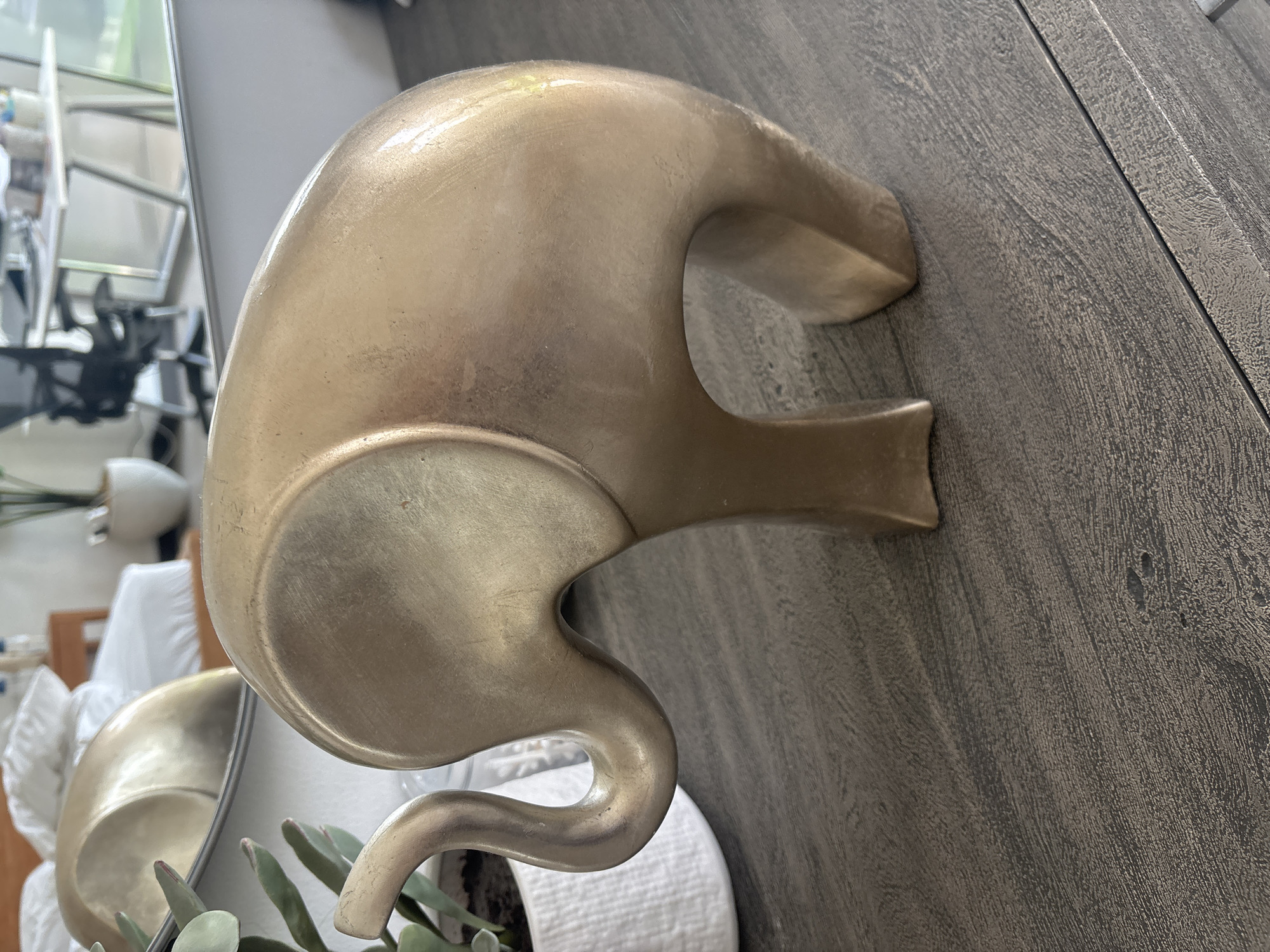 A close-up, eye-level, medium shot captures a brass elephant figurine placed on a dark, textured wooden surface. The figurine is positioned to the right of the frame, with its body facing left and its trunk curving upwards. The surface it rests on has a wood grain texture with visible lines and some indentations. In the background, a white plant pot containing greenery is partially visible to the left of the figurine. A mirror reflects a bedroom with white bedding and a wooden headboard, as well as a glimpse of an office space with a desk and chair. The overall lighting suggests an indoor environment, possibly a home or hotel room, with diffused natural light. The specific location in Dubai, United Arab Emirates, is not discernible from the visual elements in the image.
