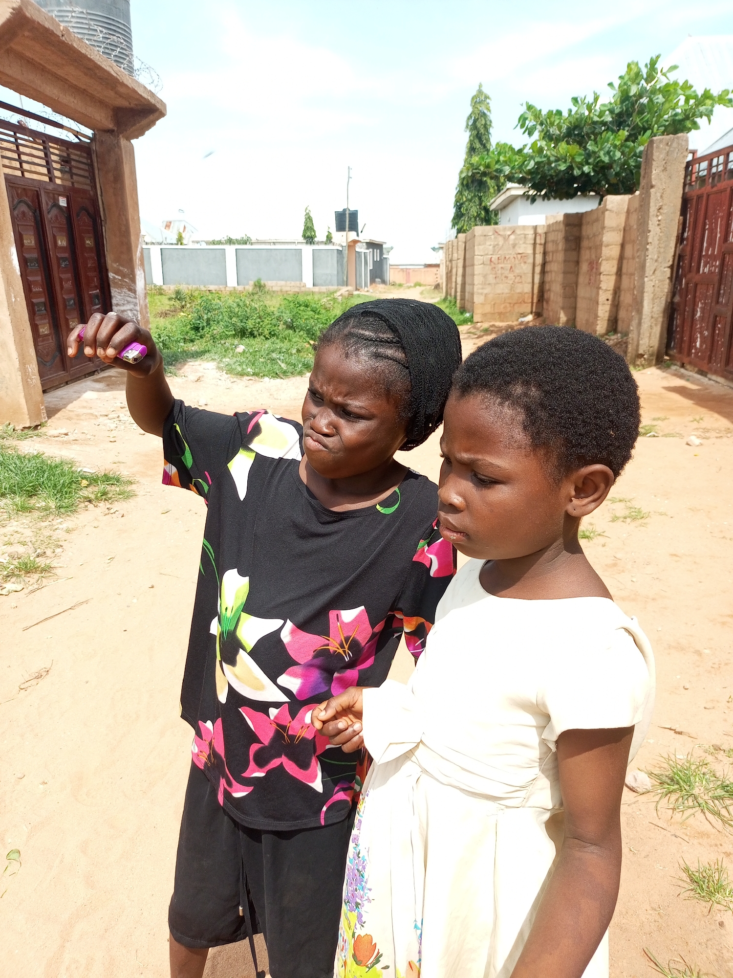 In Tempure, Nigeria, on a sunny day, two young girls are standing outdoors. The girl on the left, wearing a black t-shirt with a floral pattern and black shorts, holds a small pink object, possibly a lighter, in her outstretched hand. She is looking intently at the object. The girl on the right, wearing a light-colored dress, stands beside her, looking off to the side with a serious expression. They are in a sandy area with patches of dry grass. In the background, there are buildings, a fence, and some trees, suggesting an urban or semi-urban environment. A large water tank is visible on top of a building to the left. The overall scene depicts a moment of childhood curiosity and interaction in a Nigerian setting.