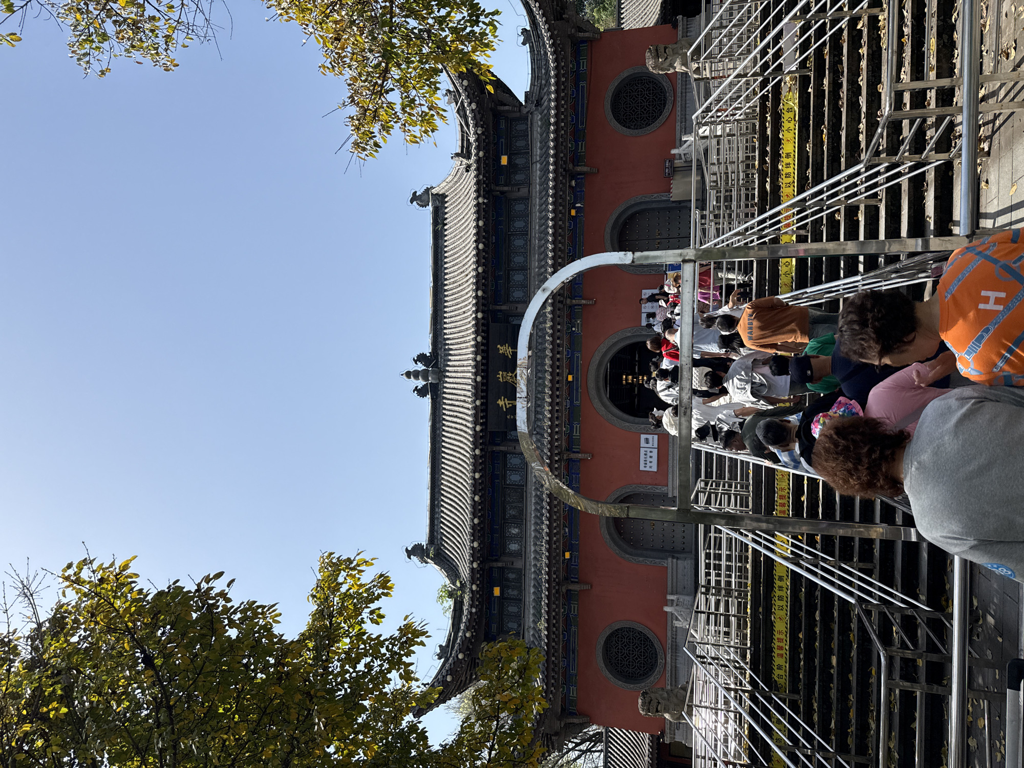 The image depicts a crowd of people on a staircase leading up to a red building with a traditional Chinese architectural style, in Jiulongpo District, China. The building has a gray tiled roof with decorative elements, a large arched entrance, and circular windows. The staircase is made of metal and has yellow caution tape. Trees with yellow leaves frame the left side of the image, and a clear blue sky is visible in the background.
