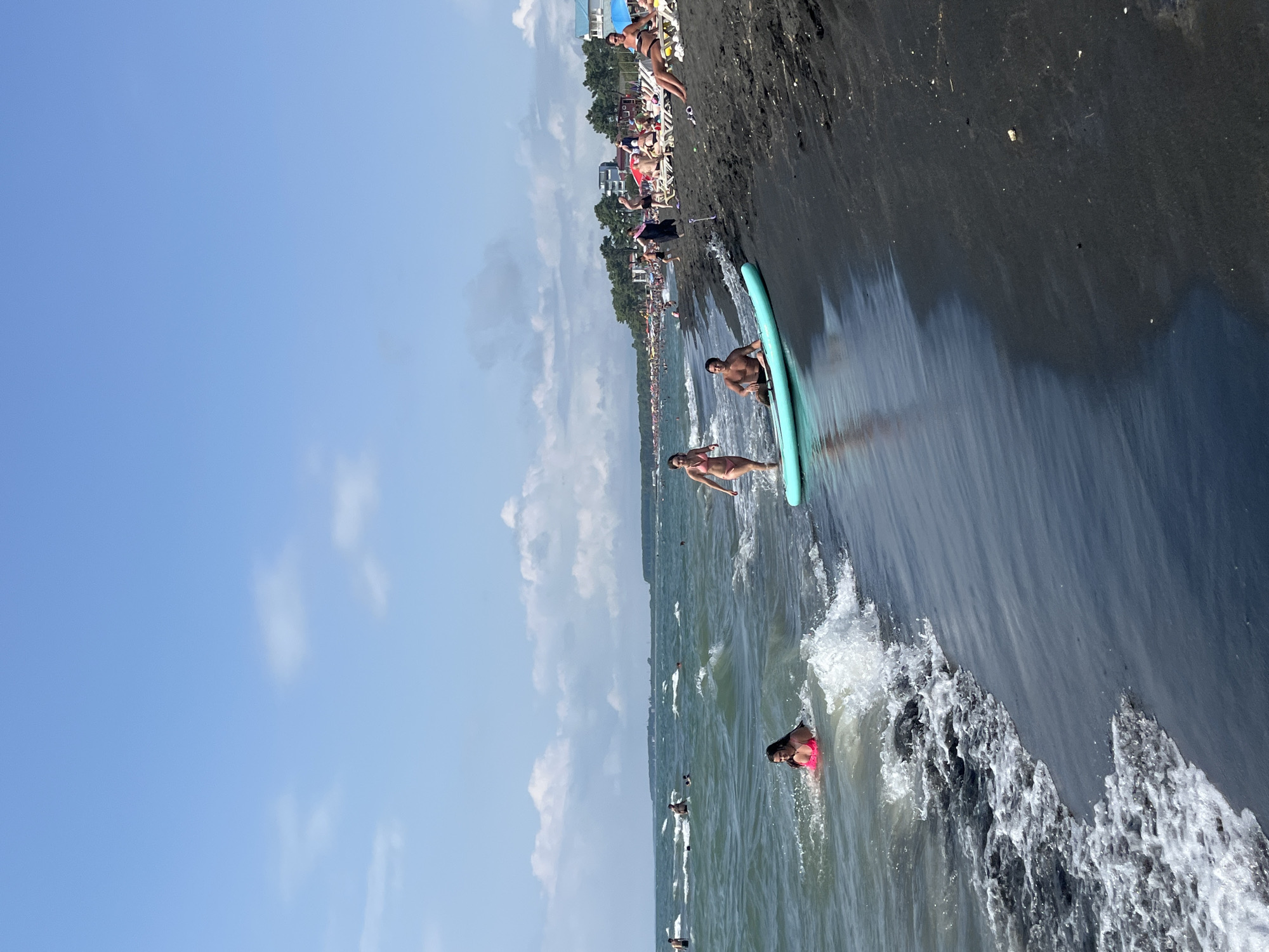 This is a photograph taken at Ureki, Georgia, featuring a black sand beach with people enjoying the water and the shore. In the foreground, the dark, wet sand meets the choppy, turquoise water. Several people are visible in the sea. One woman in a pink bikini is partially submerged, interacting with the waves. Further out, other individuals are swimming or wading.
On the shore, a light blue paddleboard is placed in the shallow water, with a man sitting on it. A woman in a pink bikini is walking out of the water towards the paddleboard. Beyond them, the beach is lined with lounge chairs occupied by sunbathers. In the background, a dense line of trees runs parallel to the coastline, with some buildings and umbrellas visible among them, indicating a resort area. The sky is a clear blue with scattered white clouds. The overall scene depicts a lively summer day at a popular beach destination.