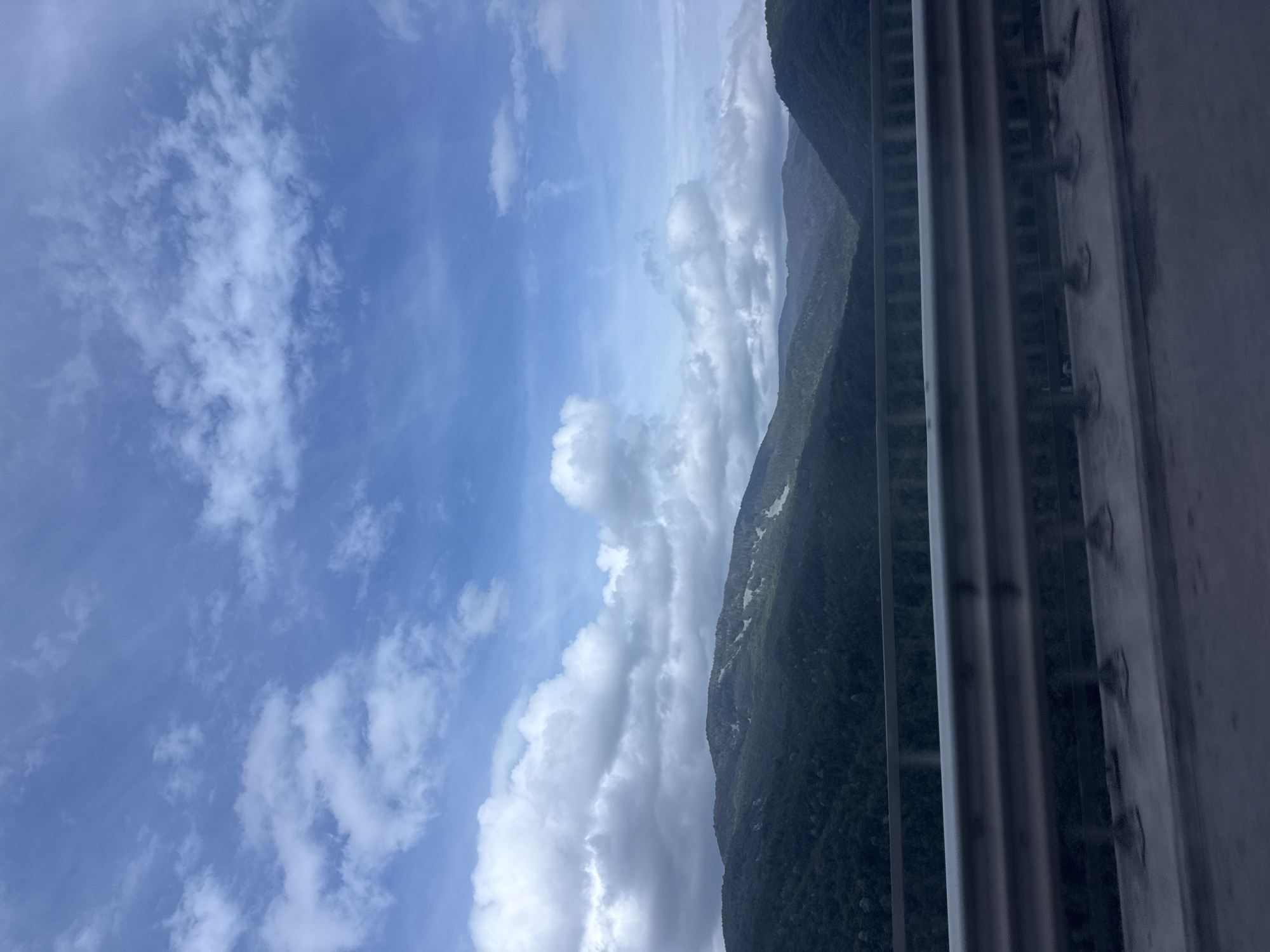 The image depicts a scenic view of rolling, tree-covered mountains under a partly cloudy sky. A metal guardrail is prominently visible in the foreground, suggesting the vantage point is from a bridge or elevated roadway. The mountains are covered in dense, dark green foliage, with some lighter patches visible on their slopes, possibly indicating clearings or rock formations. The sky is a vibrant blue, punctuated by scattered white and gray clouds, some of which are large and fluffy, while others appear more wispy. The light suggests it is daytime, with the sun likely present but partially obscured by clouds. There are no people or specific man-made structures other than the guardrail visible in the image, beyond the implication of a road. The location is identified as Churek, Bulgaria, suggesting a rural or mountainous region within that area.