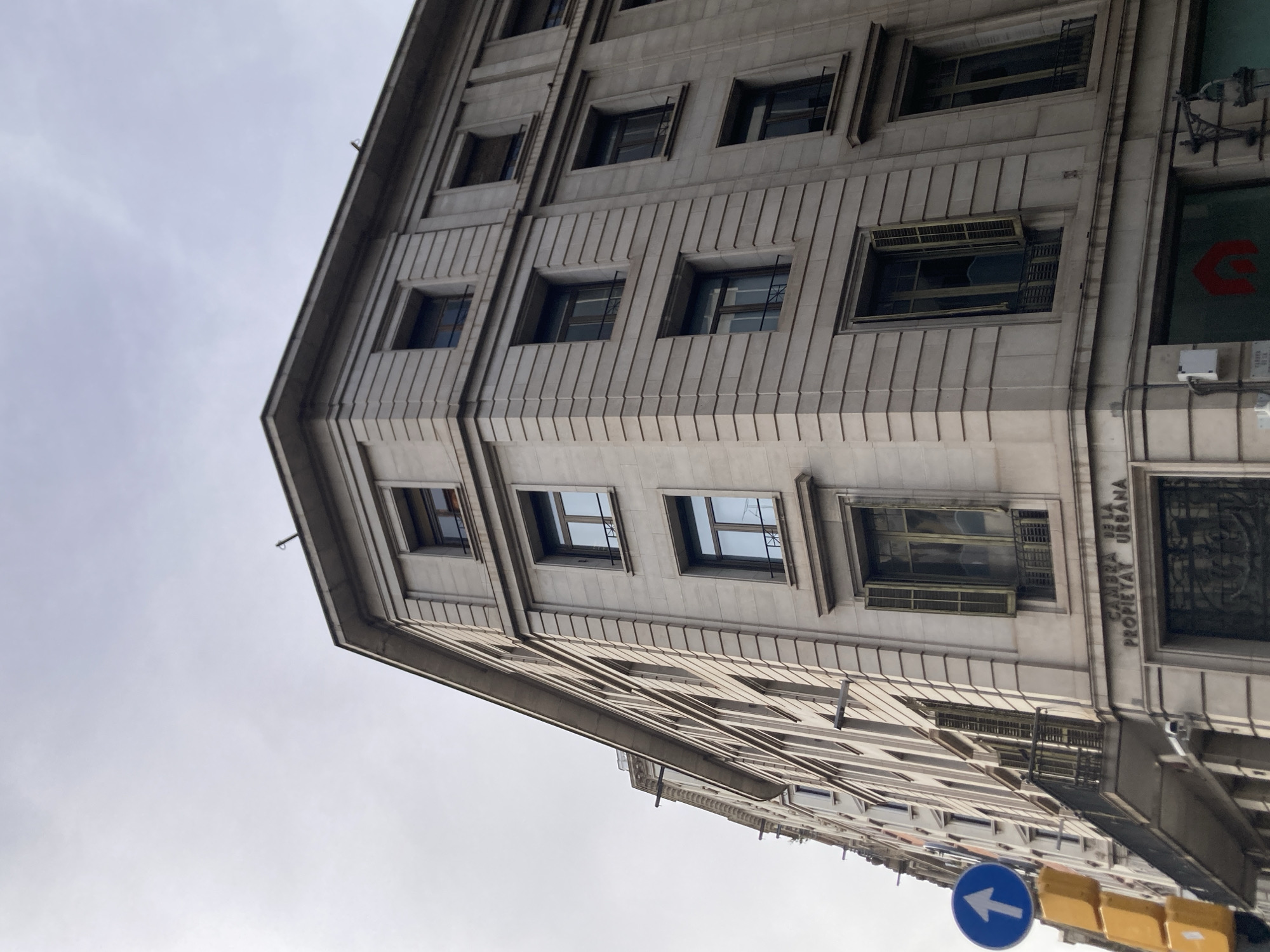 The image shows the exterior of a tall, light-colored stone building with multiple windows. The building appears to be an older architectural style with ornate detailing around the windows and along the facade. The photograph is taken from a low angle, looking upwards, and the building is angled to the right, suggesting a street corner or a unique perspective of the structure.
The sky is overcast and a pale gray, indicating a cloudy day. There are no people visible in the image. Several windows are open, revealing darker interiors or blinds. Some windows have what appear to be metal grates or security bars. On the right side of the building, some signage is visible. One sign, partially obscured, reads "CAMBRA DE LA PROPIETAT URBANA". Below this, there is a green sign with a red curved logo. Further down the street, a blue circular traffic sign with a white arrow pointing left is visible, along with yellow traffic lights.
The overall impression is of an urban environment in Barcelona, Spain, focusing on the architecture of a substantial building under a gray sky. The scene is static, with no apparent activity. The lighting is diffused, consistent with an overcast day.