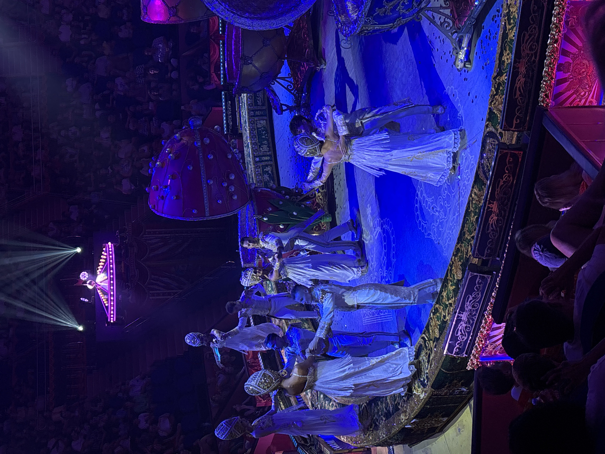 This image captures a performance at the Bolschoi State Circus in Sochi, Russia. The scene depicts a group of performers in ornate white and gold costumes, featuring elaborate headwear, dancing on a circular stage illuminated by blue and purple lights. The stage is adorned with intricate patterns and bordered by a decorative railing. In the background, large, decorative drum-like structures with red and gold embellishments are visible. Spectators are seated in the tiered audience area, with some visible in the foreground. Spotlights from above cast dramatic beams of light onto the performance space.