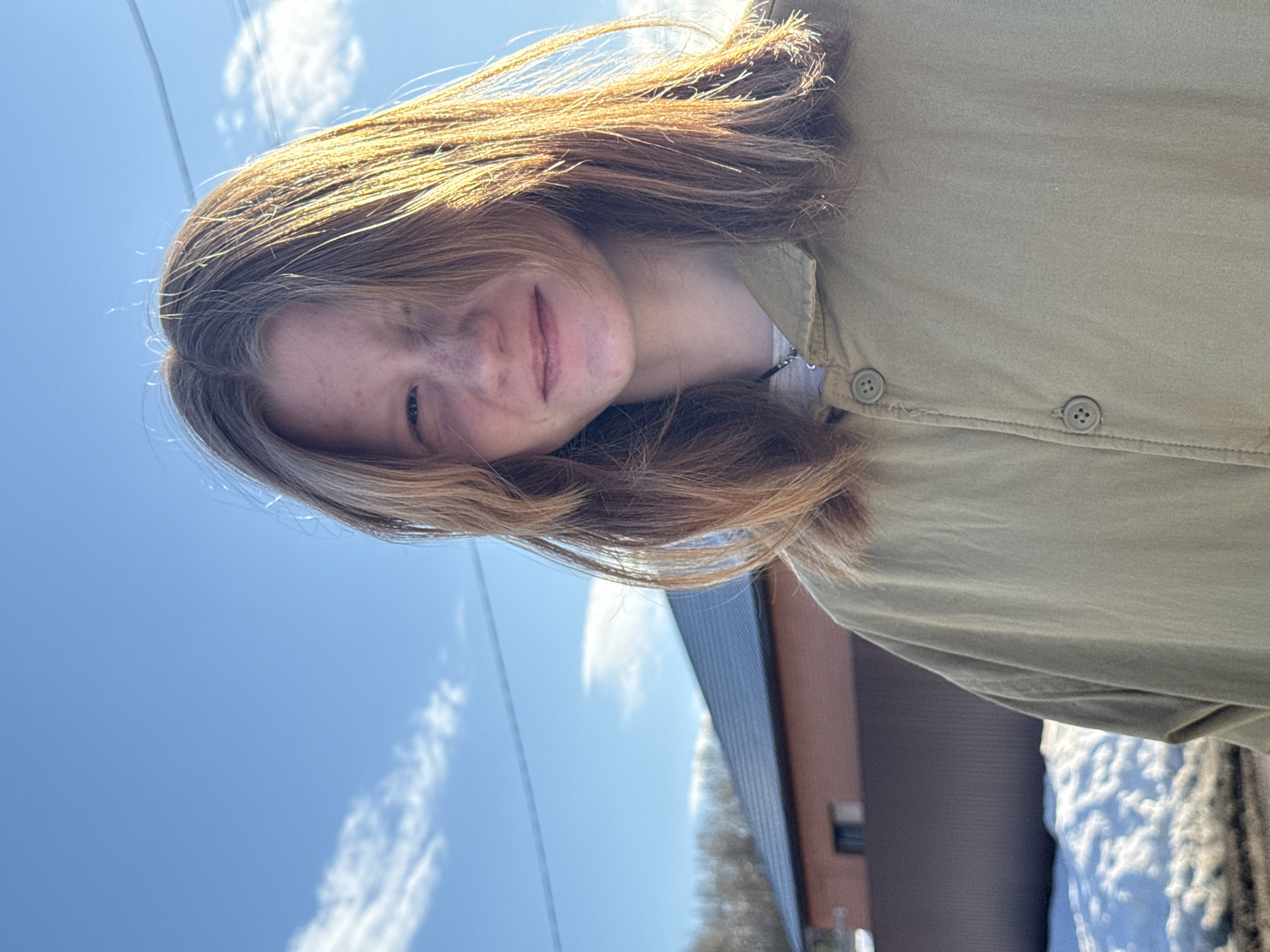 A young person with light brown hair, freckles, and wearing a khaki button-down shirt stands outside on a sunny day with a blue sky and some clouds. Behind them is a building with a brown facade, a dark gray roofline, and what appears to be a patch of snow. Power lines are visible against the sky. The location is unknown.