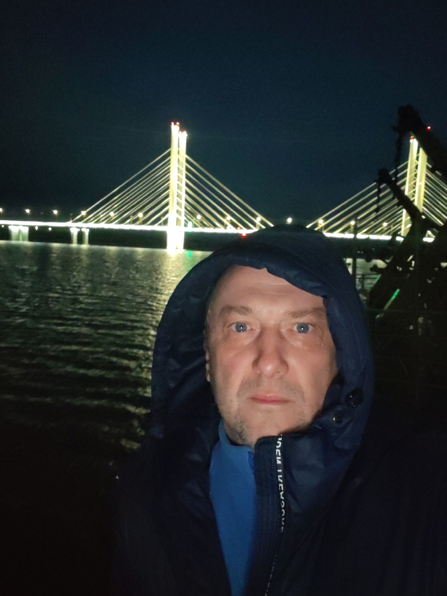 A man with light skin and blue eyes, wearing a dark blue hooded jacket and a blue shirt underneath, is taking a selfie at night. The background features a body of water, and a cable-stayed bridge illuminated with white and yellow lights. The man is centered in the foreground with the bridge centered in the background. The surrounding environment is dark. The bridge is identified as the Astana Bridge, located in the city of Nur-Sultan, Kazakhstan.