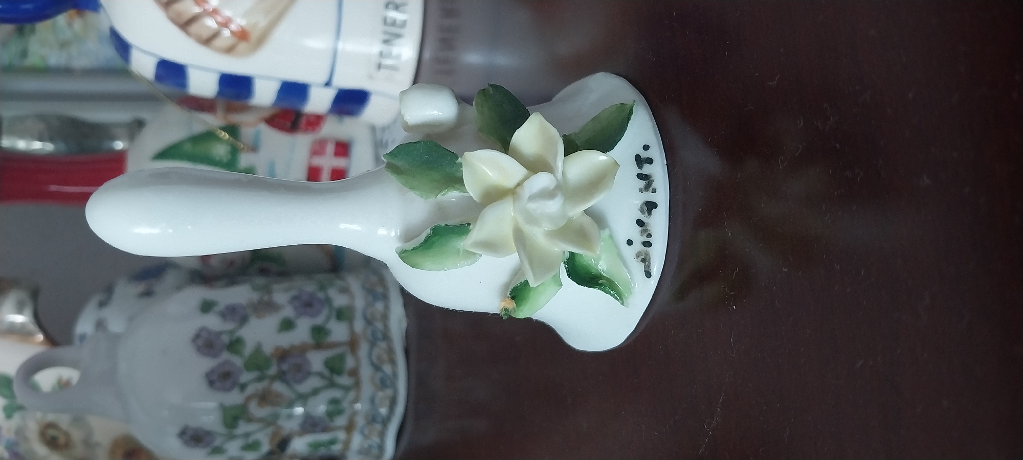 A close-up shot focuses on a white ceramic bell decorated with a delicate pale yellow flower and green leaves. The bell is positioned on a dark brown, reflective surface. In the background, out of focus, are other decorative bells, including one with a blue and white striped pattern and another adorned with floral motifs. The overall scene suggests a collection of decorative items, likely displayed on a shelf or table. There is no indication of a location in Moscow, Russia, or any people present.