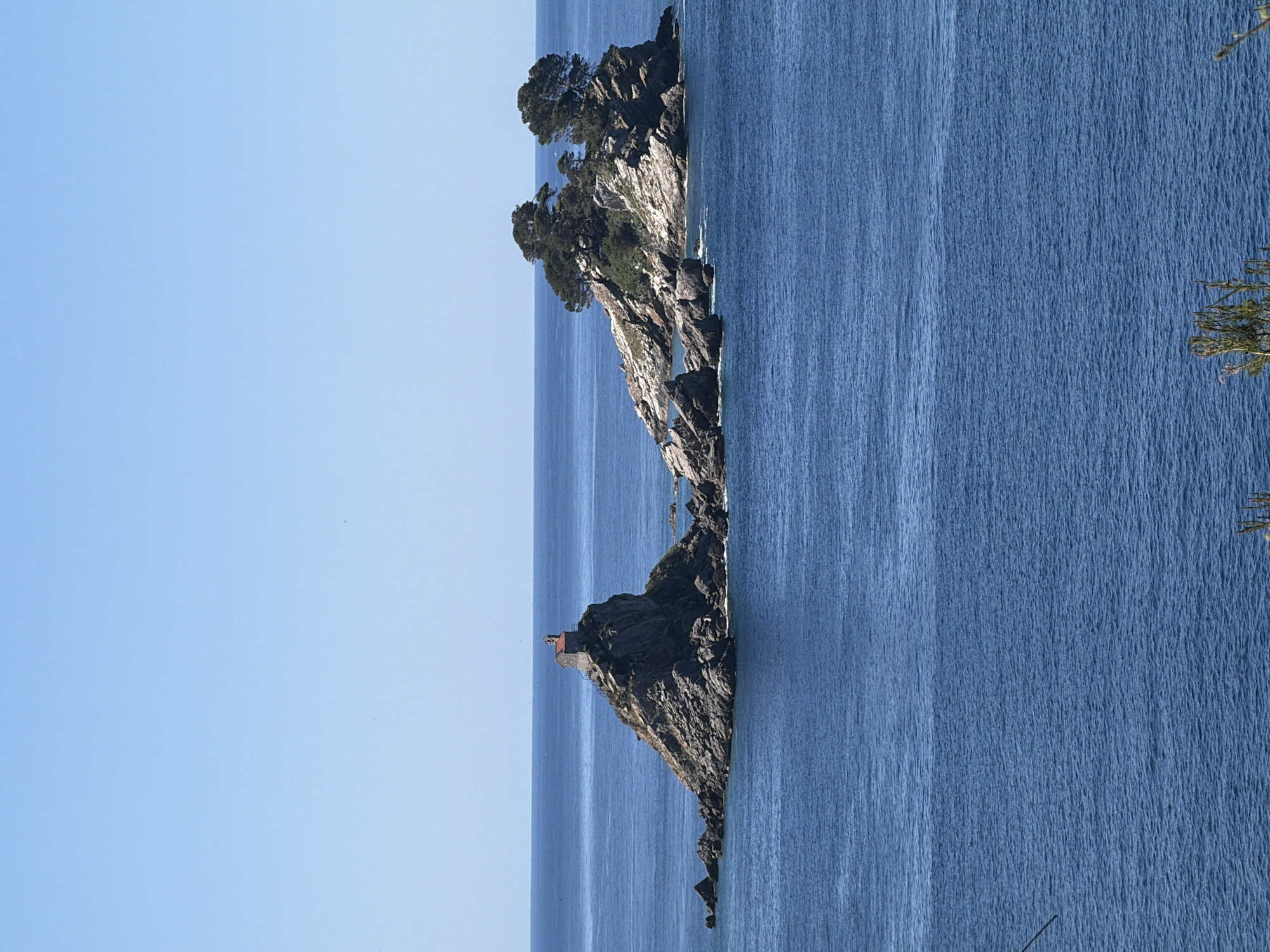 The image shows a rocky coastline along the sea, likely near Petrovac centar, Montenegro. There are two prominent rock formations with vegetation, one featuring a small structure atop it. The water is a deep blue with visible texture, and the sky is clear and light blue.