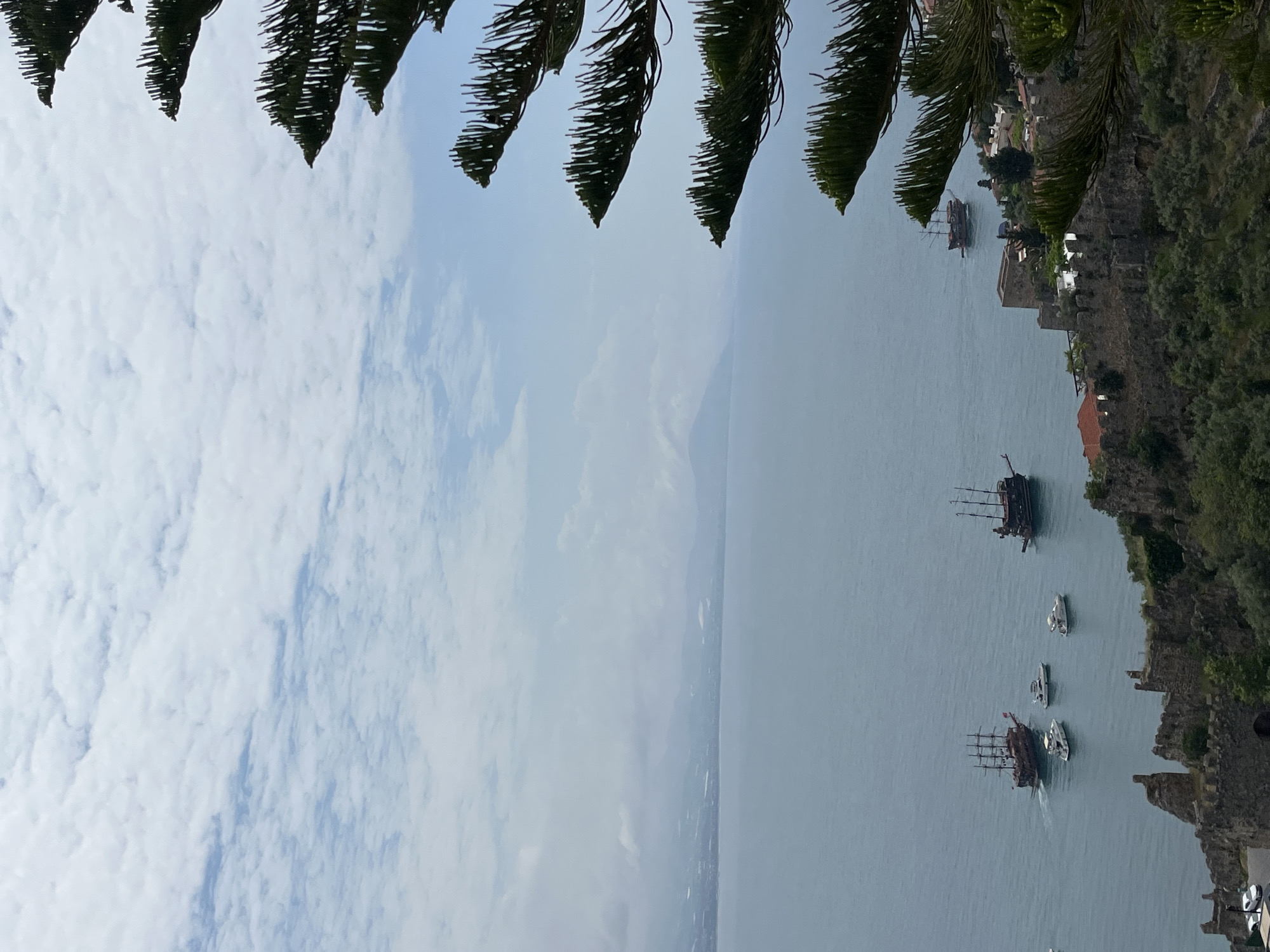 The image shows a view of the sea and coastline in Alanya, Turkey, taken from an elevated position. The scene is partially framed by pine tree branches in the foreground. Several boats are visible on the water, including at least two distinct galleon-style pirate ships, and several smaller yachts or motorboats. The coastline is rocky and verdant with visible historical buildings. The sky is overcast with a mixture of clouds.