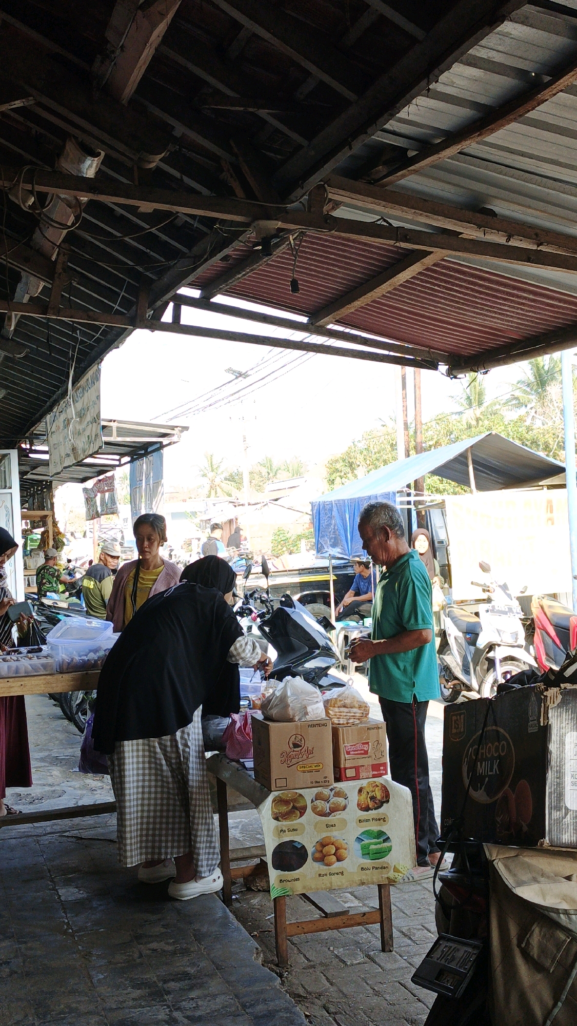 The image depicts an outdoor market scene in Cibenda, Indonesia. The foreground shows a stall with various food items displayed on a table and in boxes. A person wearing a checkered skirt and black headscarf is interacting with a vendor, a man in a green shirt, over the table. The vendor holds a small item in his hands, possibly money or a product. Behind them, a motorcycle is parked.
In the background, several other people are visible, some standing and some sitting. One woman in a yellow shirt and headscarf stands near the stall. Further back, a blue tarp covers some structures, and more motorcycles are parked. The market appears to be covered by a rustic wooden and corrugated metal roof. Palm trees and other vegetation are visible in the distance, suggesting a warm climate. The overall atmosphere is that of a busy, open-air marketplace.