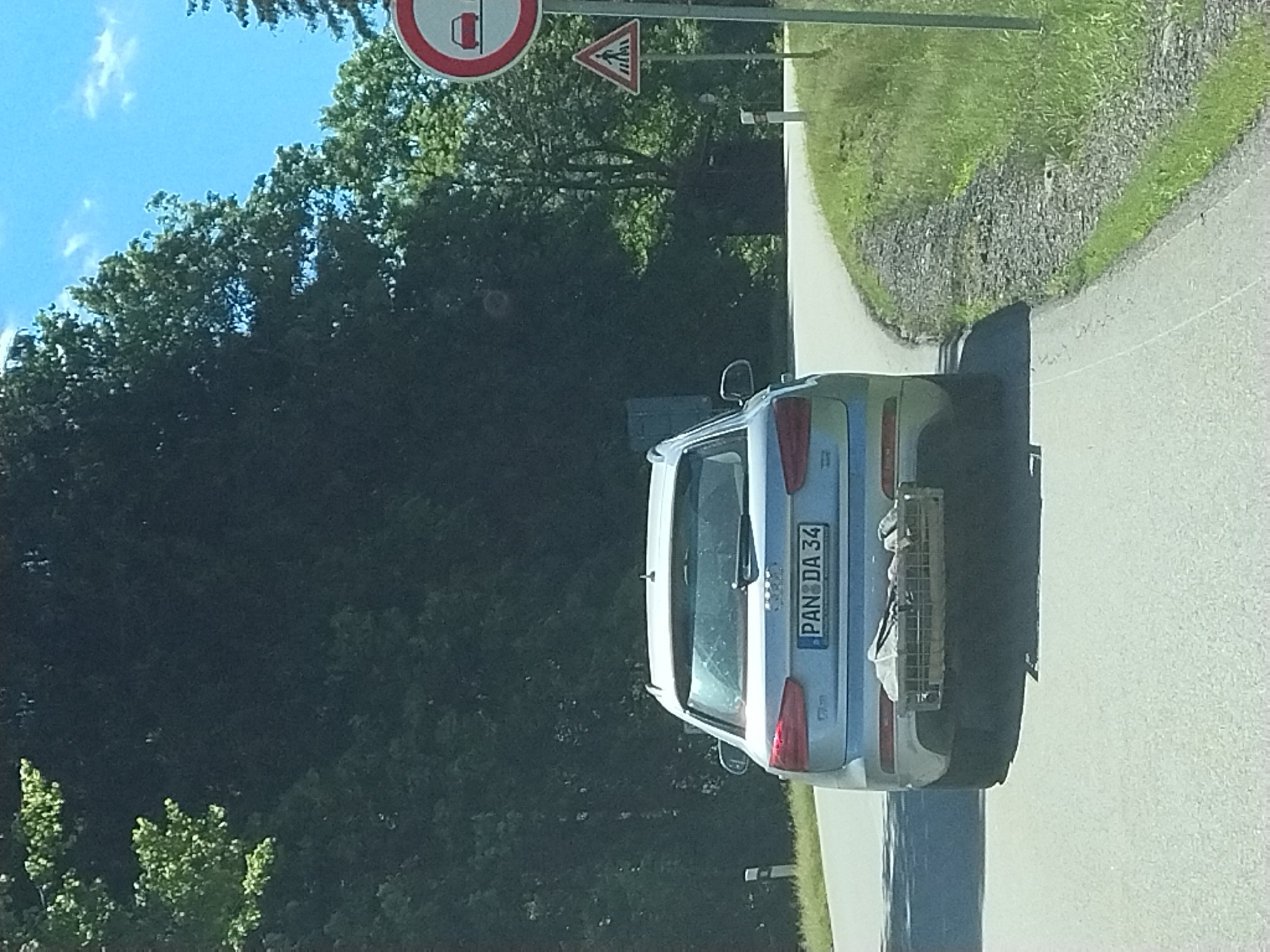A silver Audi station wagon is parked on the side of a road, with its rear facing the viewer. The car has a license plate that reads "PAN DA 34". A metal rack is attached to the rear of the car, holding some items. The scene appears to be in a rural area, with a tree-lined road and grassy slopes. The sky is blue with some scattered clouds, suggesting it is daytime and the weather is fair. The overall atmosphere is peaceful and serene.
The image is taken from a slightly elevated angle, looking down at the car. The car is positioned at an angle to the road, as if it has just been parked or is about to be driven away. The focus is on the car and its surroundings, with the background slightly blurred. The sunlight casts shadows on the road and the car, indicating a bright day.
The presence of road signs in the background, though partially obscured, suggests that this is a public road. The trees and greenery indicate a natural environment, likely a forest or park. The road itself appears to be asphalted.
The image seems to be a snapshot taken during a road trip or a casual stop in a scenic location. There are no people visible in the image, but the car implies the presence of occupants. The overall impression is one of a pleasant drive through the countryside.