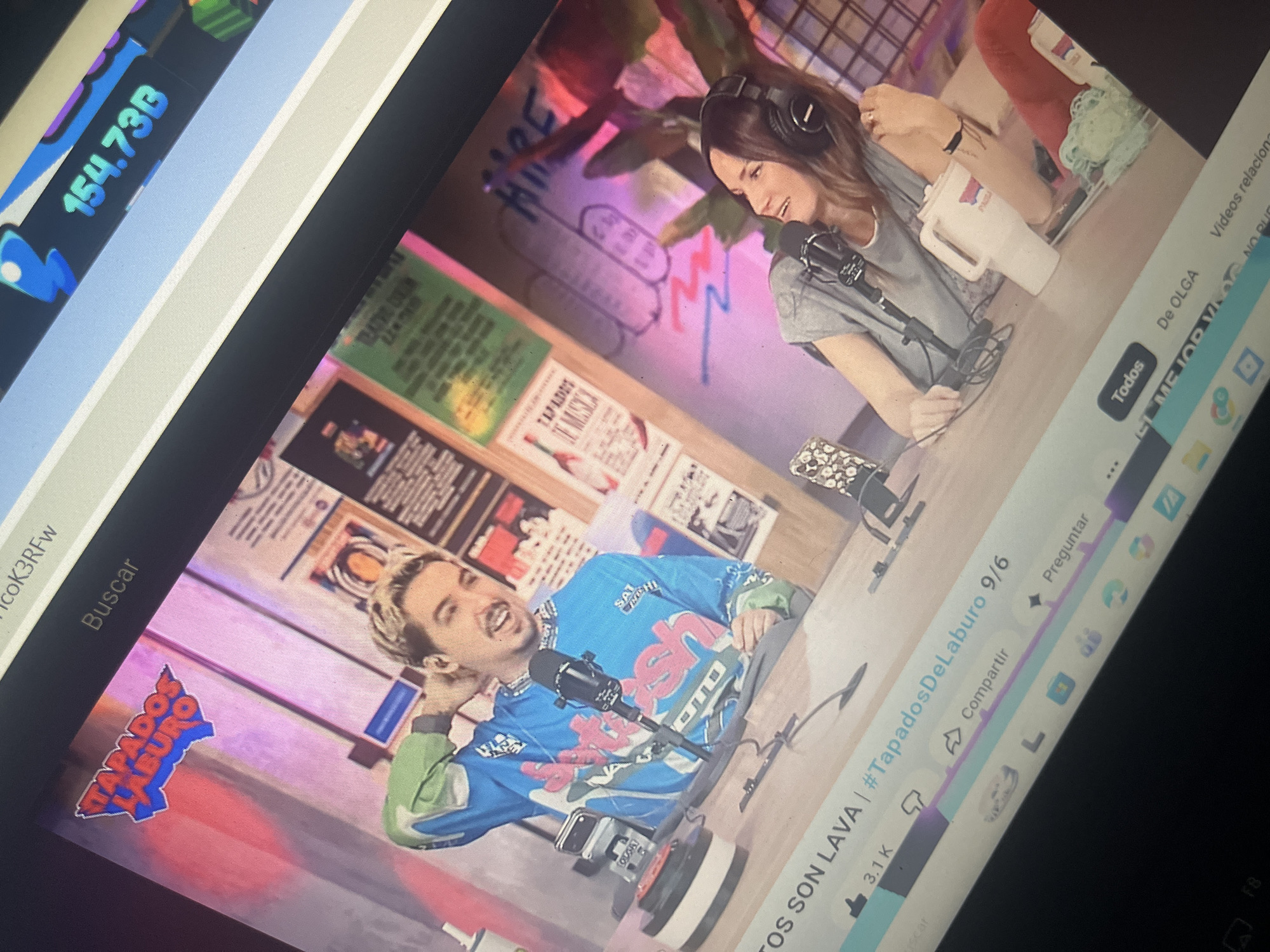 The image depicts a screen displaying a video stream, likely from a platform like YouTube. On the screen, two people are visible, seated at a table in what appears to be a studio setting. The person on the left is a male with blond hair, wearing a blue and green sweatshirt with a graphic design. He is looking at the camera and has his hand on the back of his neck. In front of him is a microphone and a red button. The person on the right is a female with long brown hair, wearing headphones and a gray t-shirt. She is looking to her right, with a microphone in front of her. A white pitcher is on the table near her. The background features posters on the wall and some plants.
The bottom portion of the screen shows interface elements of the video player, including a title that seems to read "OS SON LAVA | #TapadosDeLaburo 9/6", likes, share options, and comments.
Regarding the location of Santa Ana, Venezuela, there is no visual information in the provided image that directly or indirectly indicates this specific location. The scene is a studio environment, and no landmarks or context suggesting Santa Ana are present.