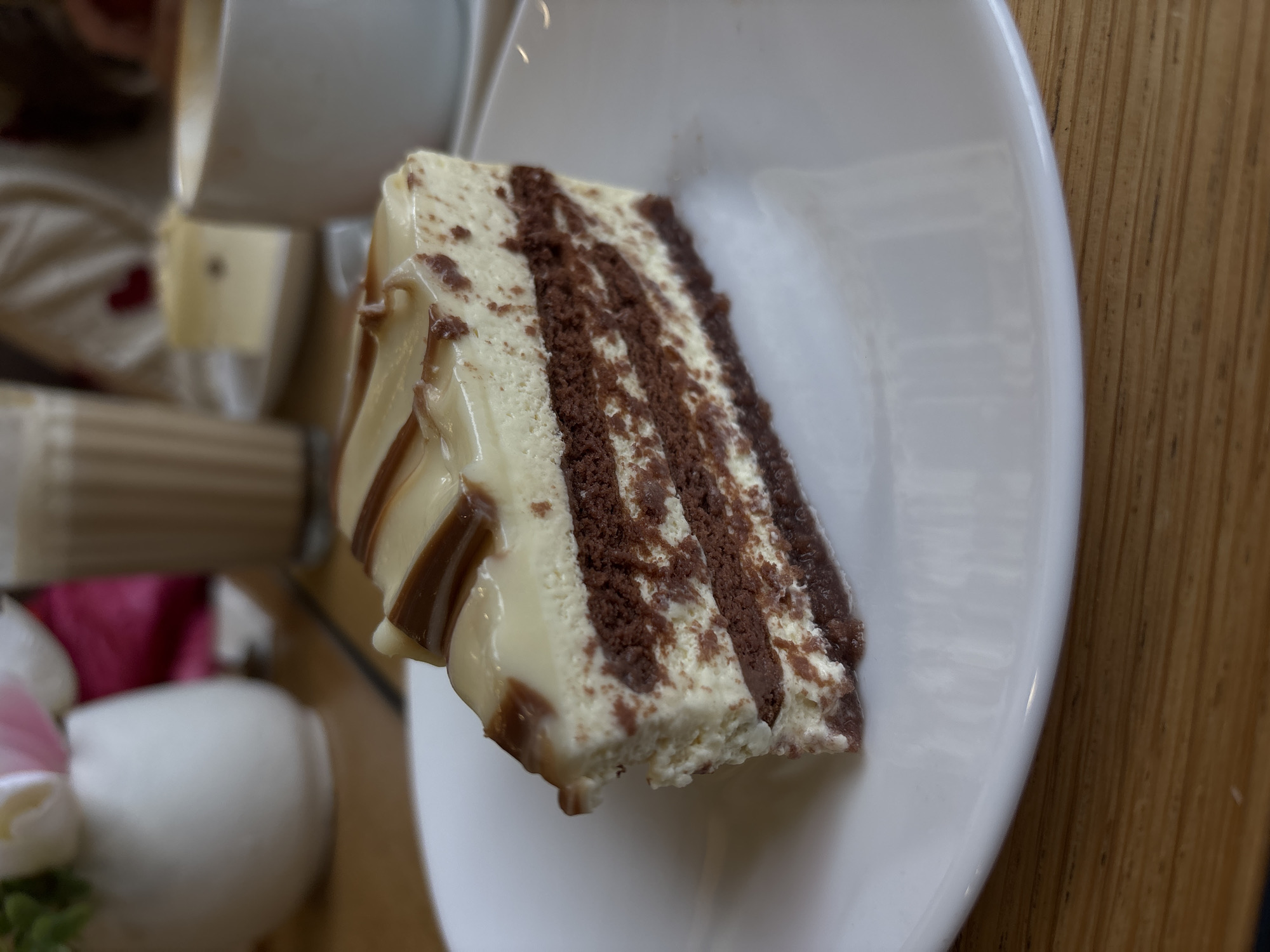A slice of layered cake sits on a white plate. The cake features alternating layers of white cream and brown cake, with a white glaze drizzled with brown sauce on top. To the left of the plate, partially visible, are other dishes and decorative elements, including white porcelain pieces and a floral arrangement. The background includes a light-colored wooden surface. The scene likely takes place indoors, in an eatery. Specific location in Varna, Bulgaria cannot be ascertained from the image.
