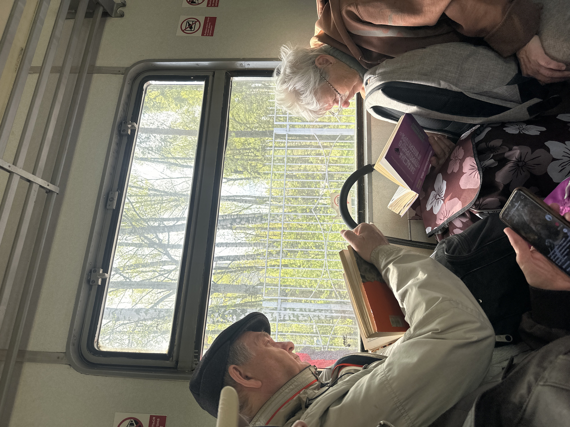 An image taken in a transit setting, possibly a train or bus, shows two older individuals occupied with reading different books. The man is in the lower section of the shot, wearing a cap, and holding a book. The woman is in the upper section, wearing glasses, and looking at a book. In the bottom right, a cell phone is being used by a third individual. The background is a window that shows a tree. There are signs near the top of the window, as well as signs on the left and bottom of the window, with Russian writing. The window has gray siding.